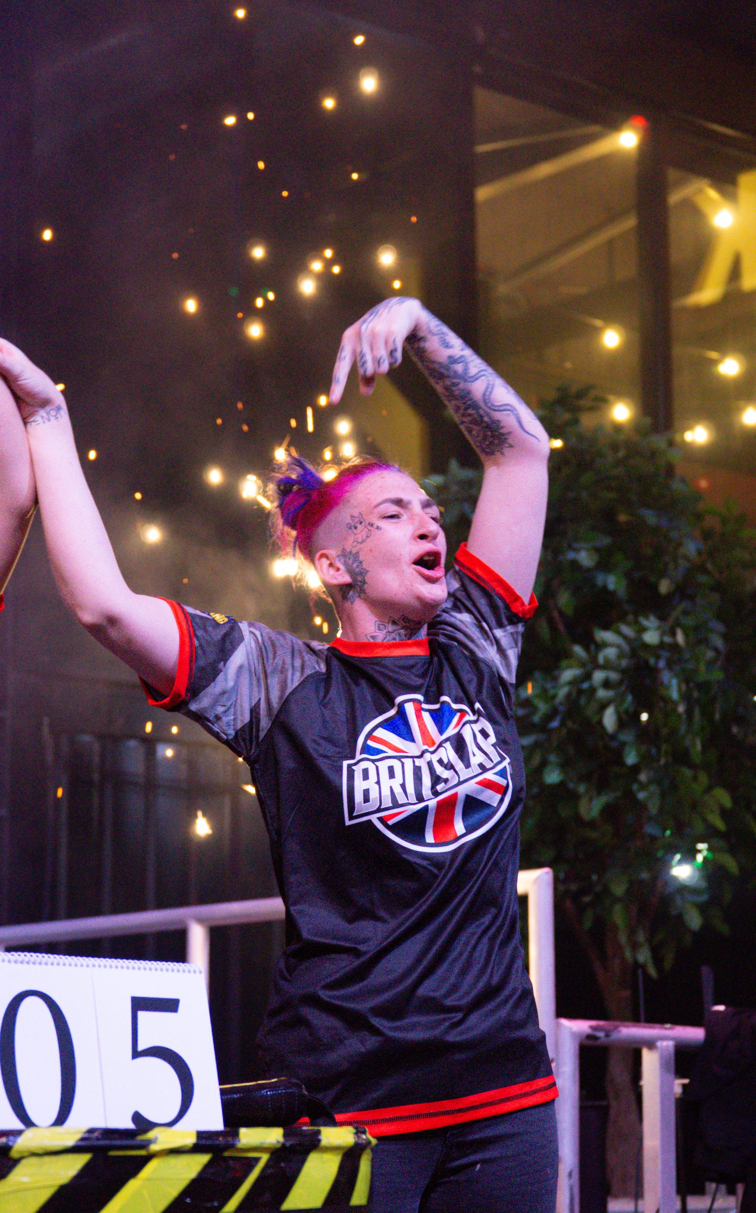 Tattooed athlete in British flag jersey celebrates with raised arms at night competition venue with string lights overhead.