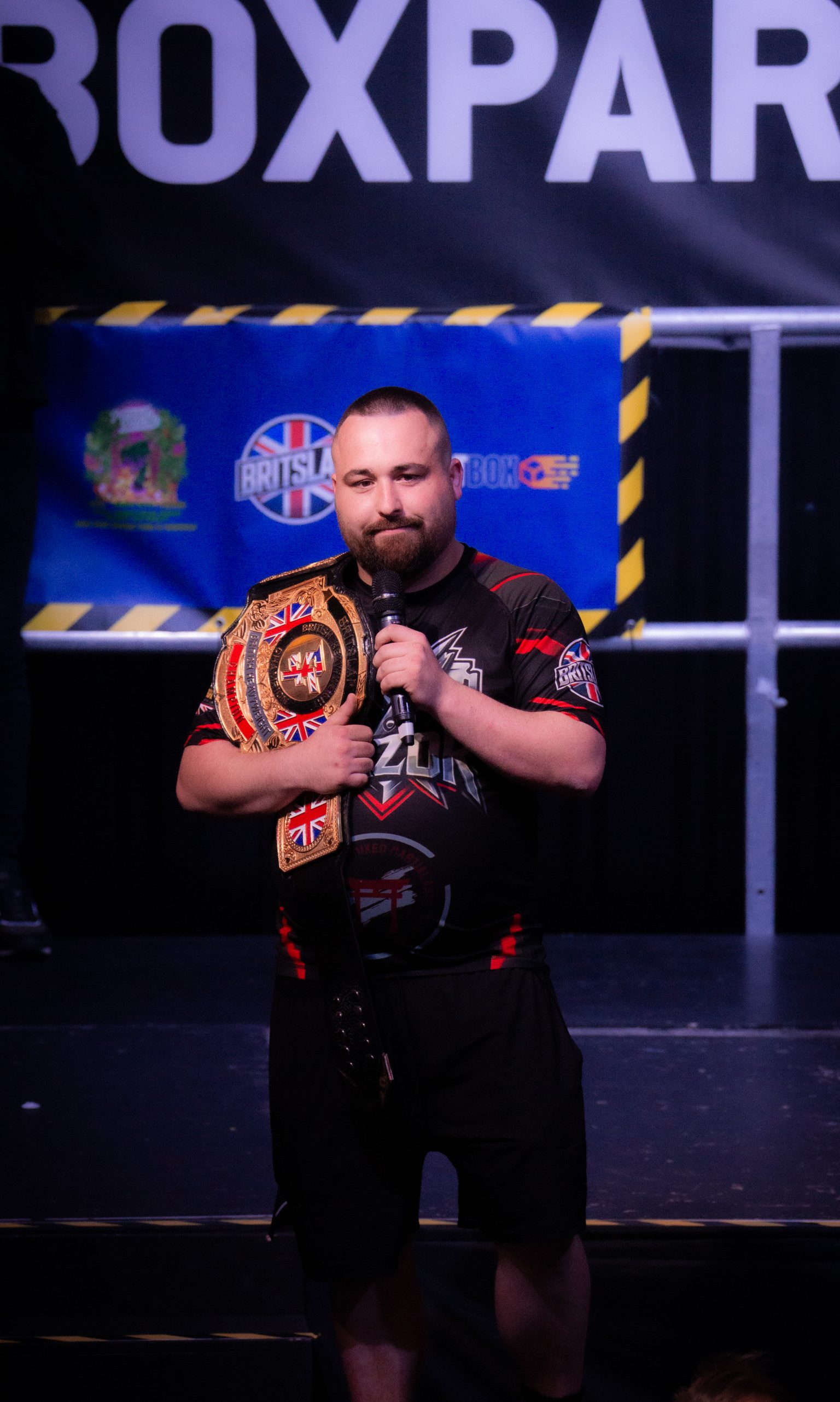 Bearded wrestler in black gear with Union Jack elements holds championship belt, standing in wrestling ring with blue ropes.