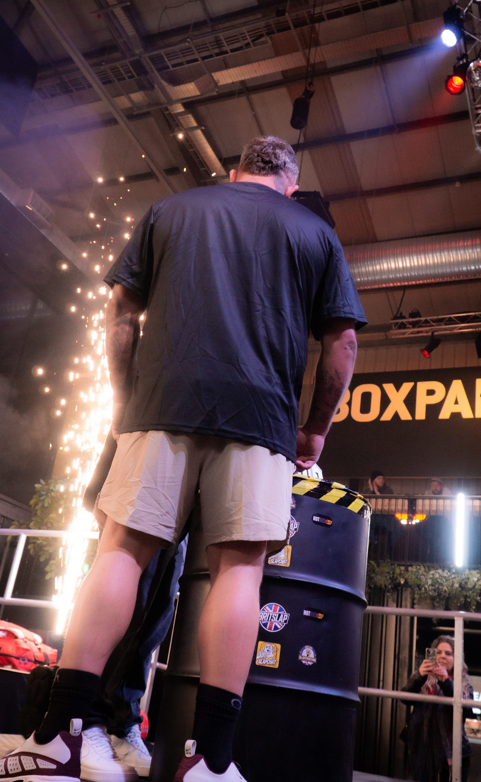 Man in navy shirt and white shorts stands next to black equipment with bright sparks shooting upward in industrial venue.