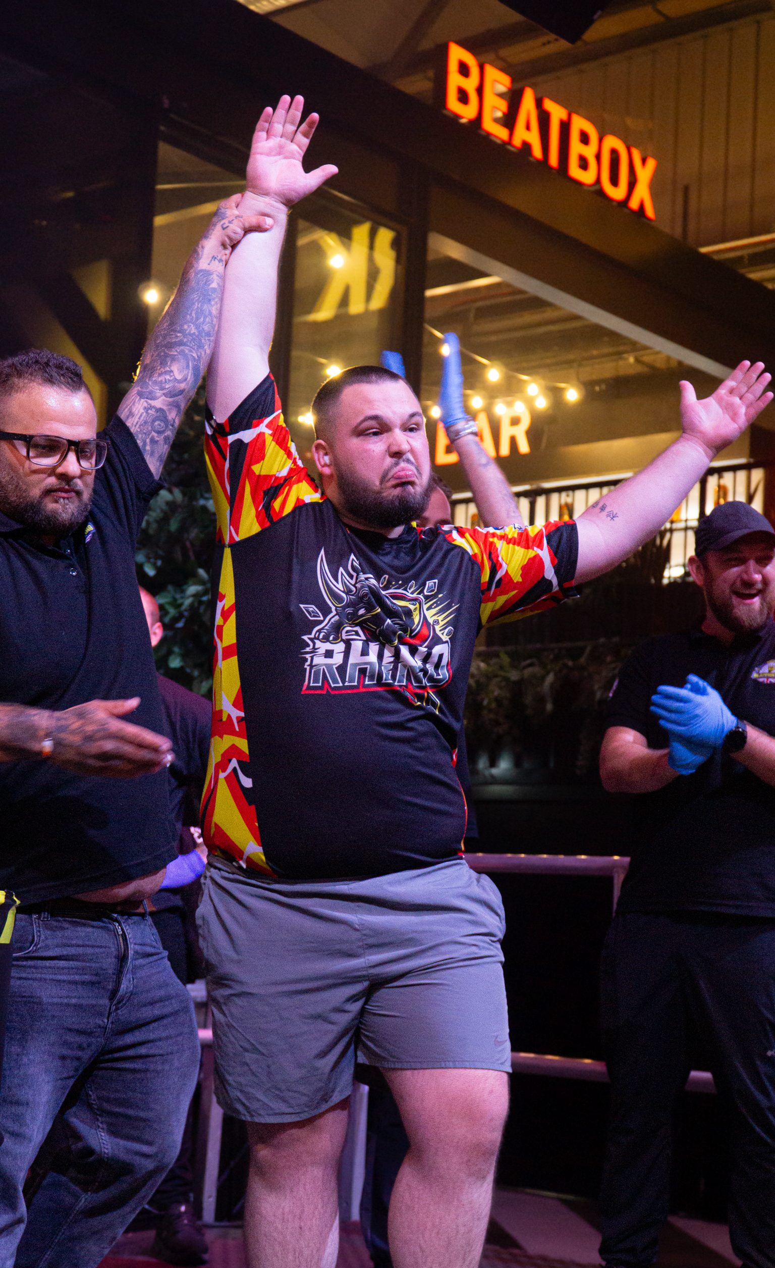 Man in black t-shirt and shorts celebrates with arms raised, surrounded by crowd. Orange "BEATBOX" sign visible above in background.