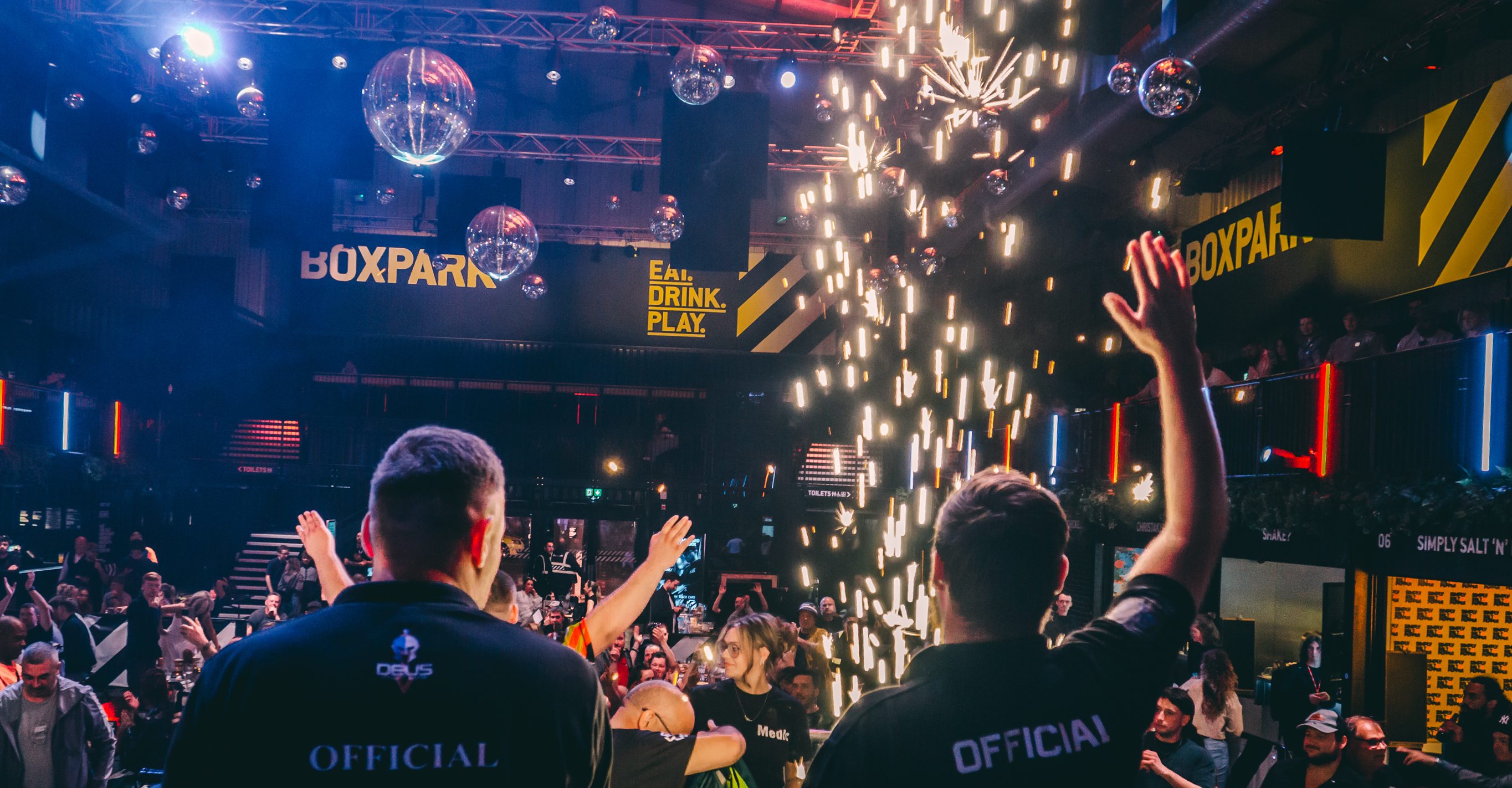 Two officials with raised arms face crowd as sparks cascade from ceiling in dimly lit venue with blue and orange lighting.