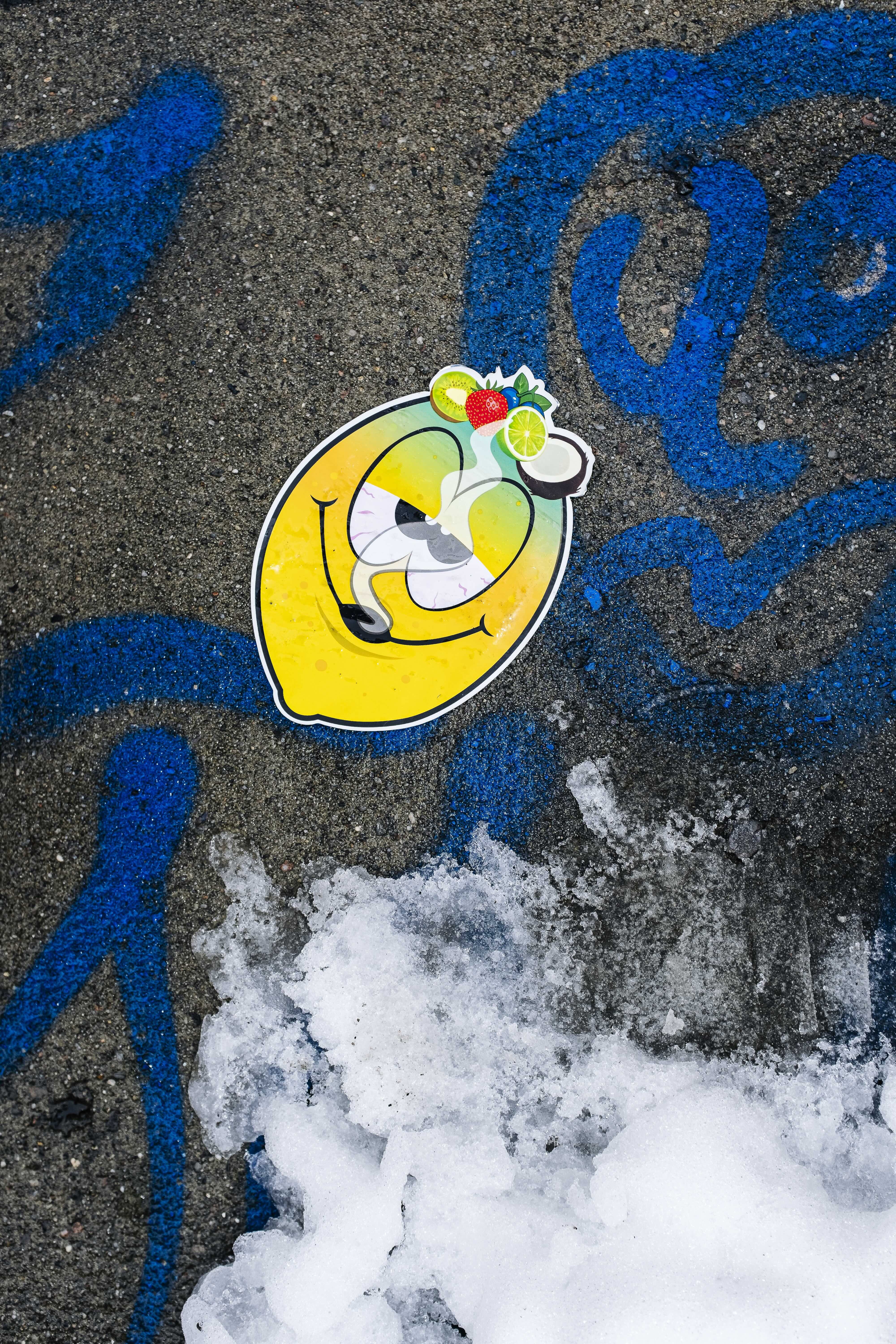 Bright yellow smiley face image with fruit accessories on snowy pavement, with blue swirls around.