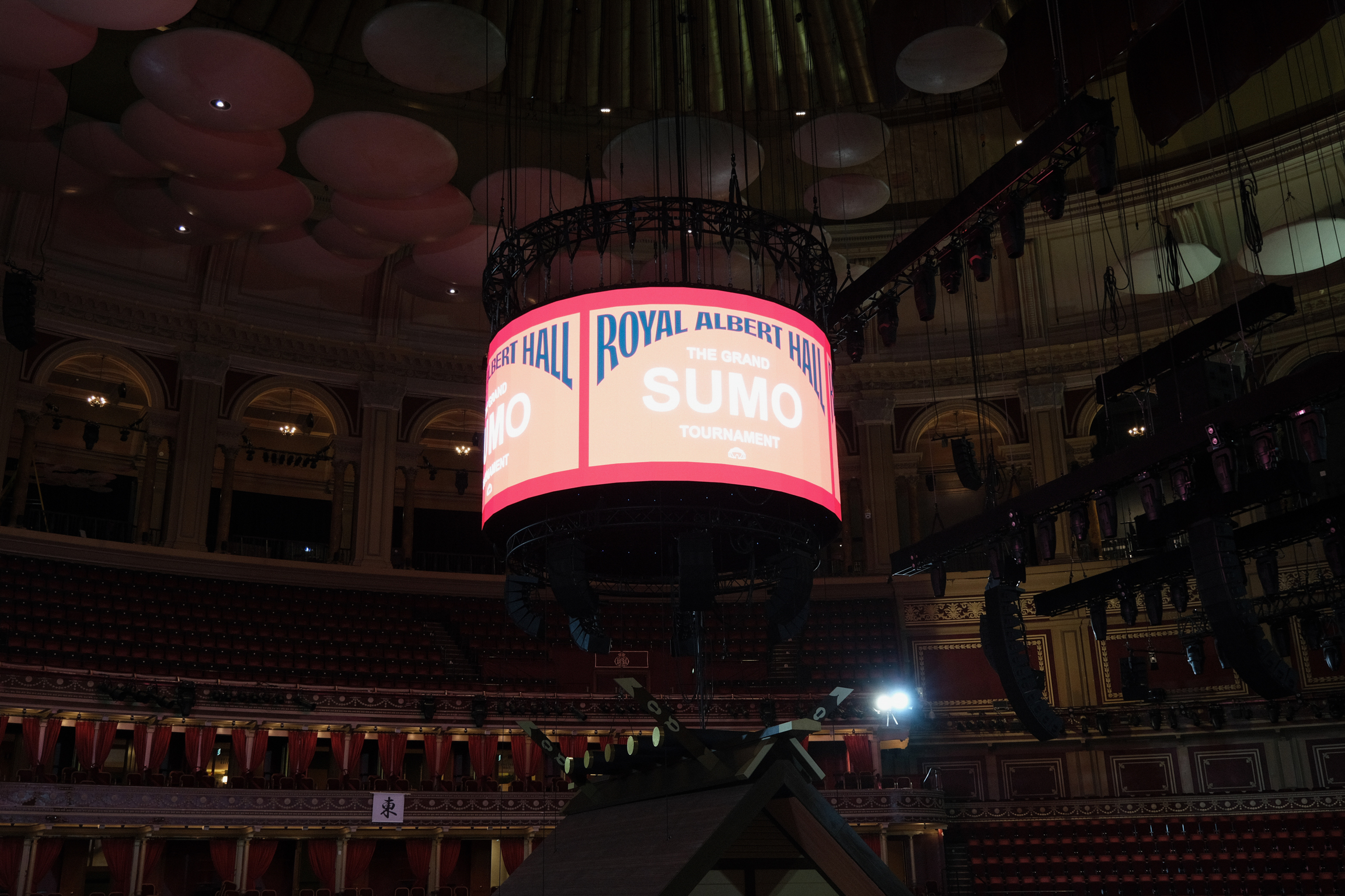 Large circular LED screen displaying "Royal Albert Hall" text suspended above red velvet theatre seating in ornate Victorian venue interior.