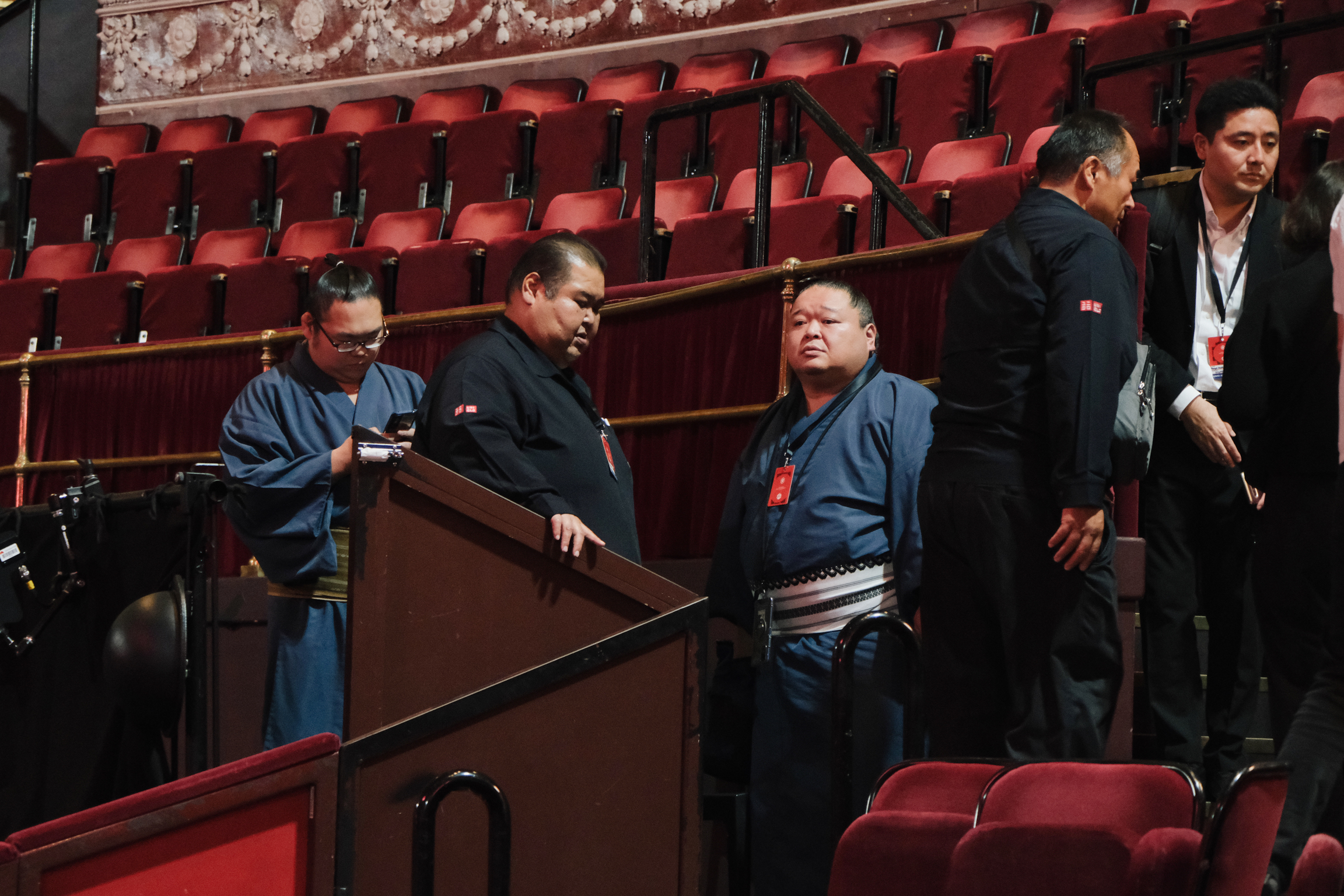 Five men in dark suits gathered around wooden barrier in theatre with red velvet seats and ornate ceiling details.