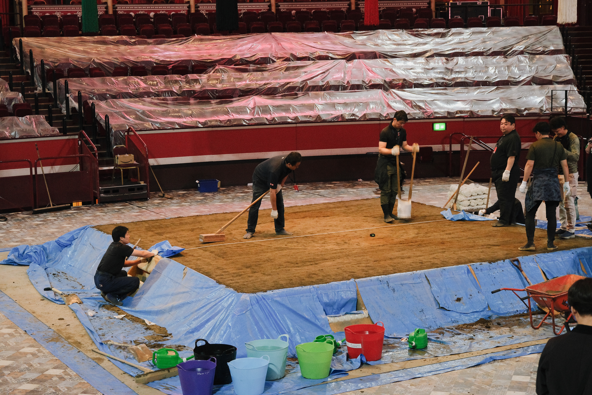 Workers laying wooden flooring on blue protective sheeting in empty stadium with red seating visible in background.