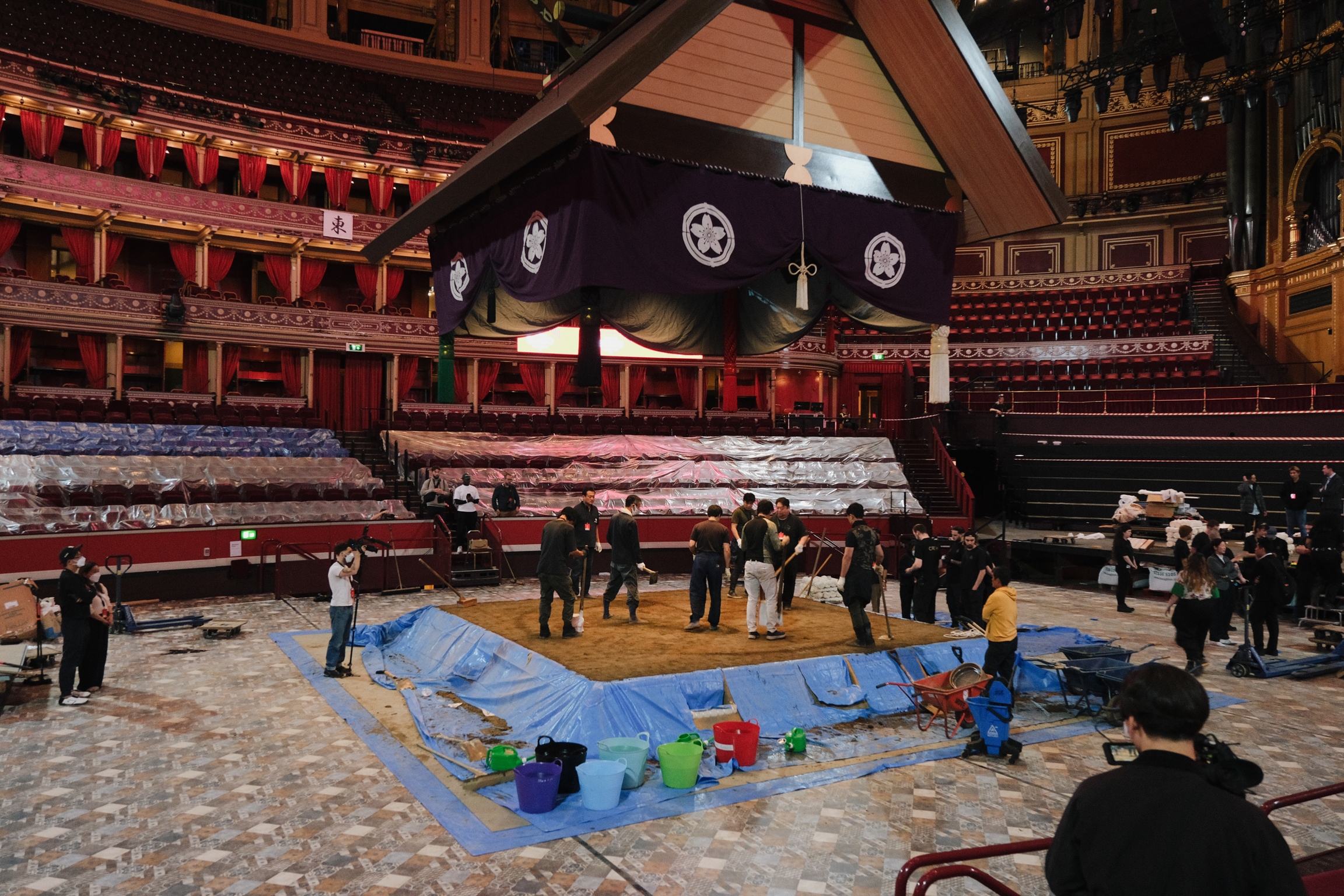 Large black inflatable structure suspended above Royal Albert Hall arena floor with crew members working on blue tarpaulin below.