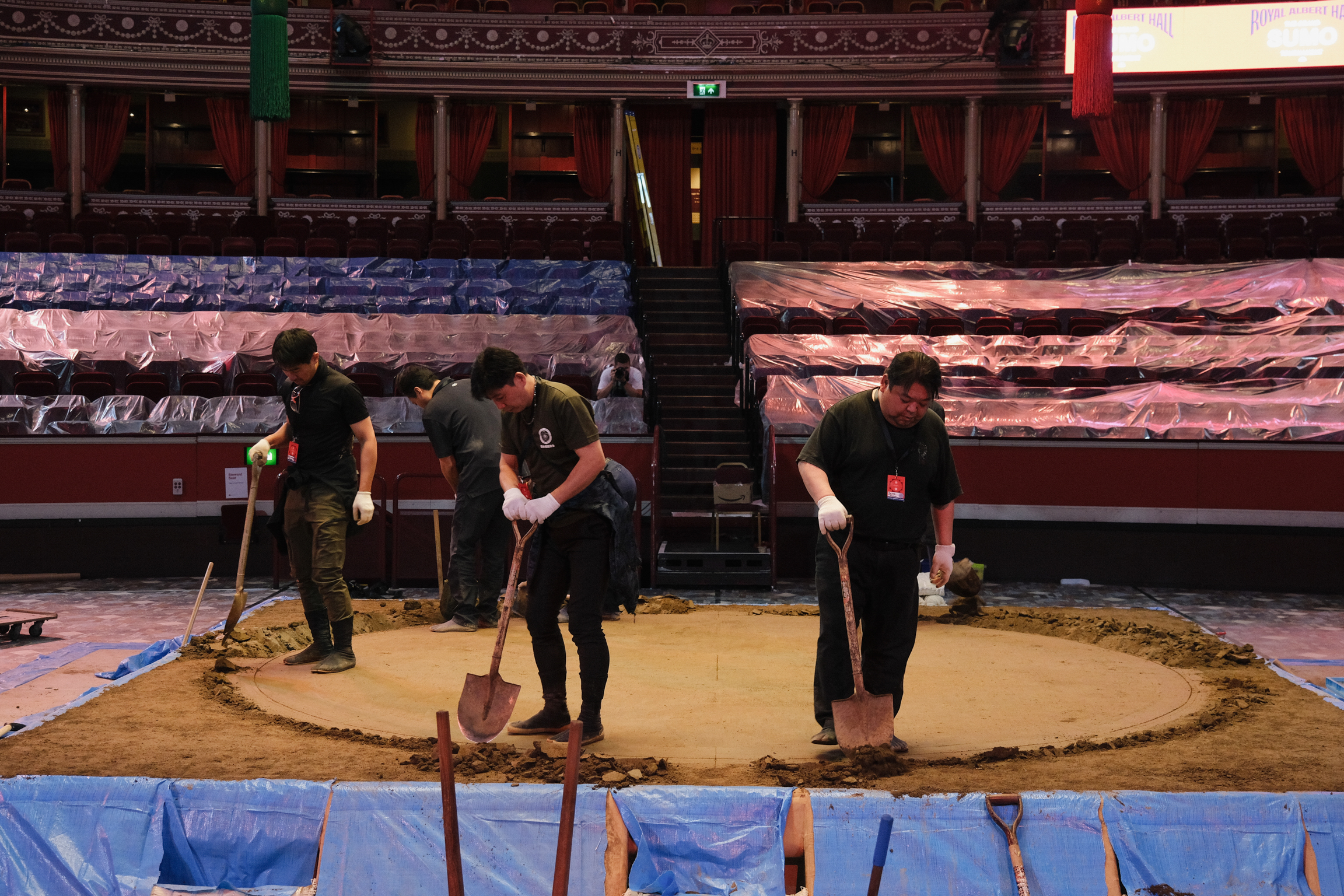 Three workers with brooms sweeping sand on a large tarpaulin inside an empty stadium with red and blue seating.