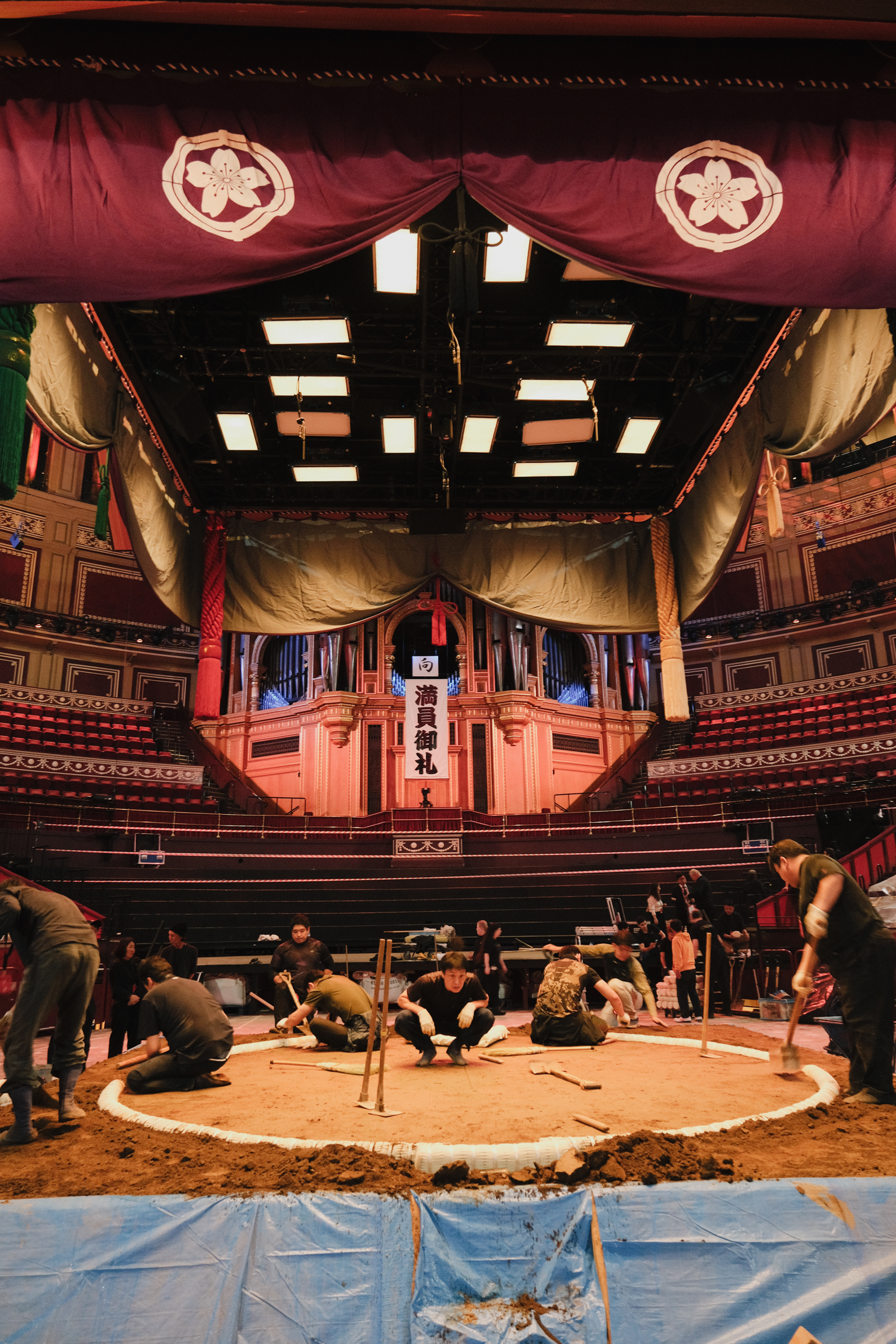 Traditional sumo wrestling ring with spectators seated around circular sandy arena, ornate wooden architecture, red fabric ceiling.