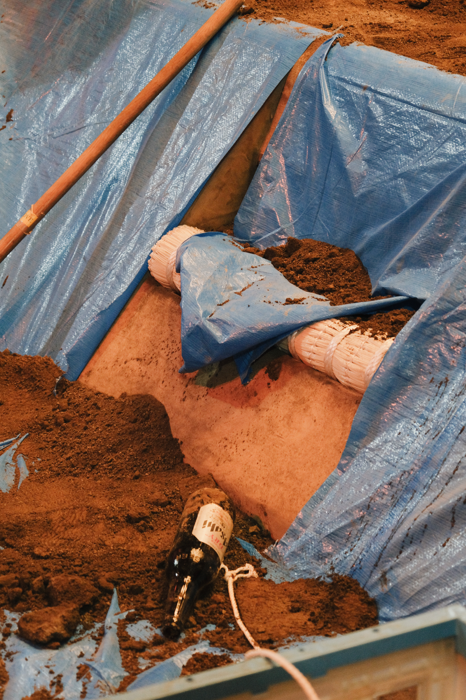 Blue plastic sheeting covers orange-brown soil with exposed patches. White cylindrical objects and electrical components visible beneath torn plastic covering.