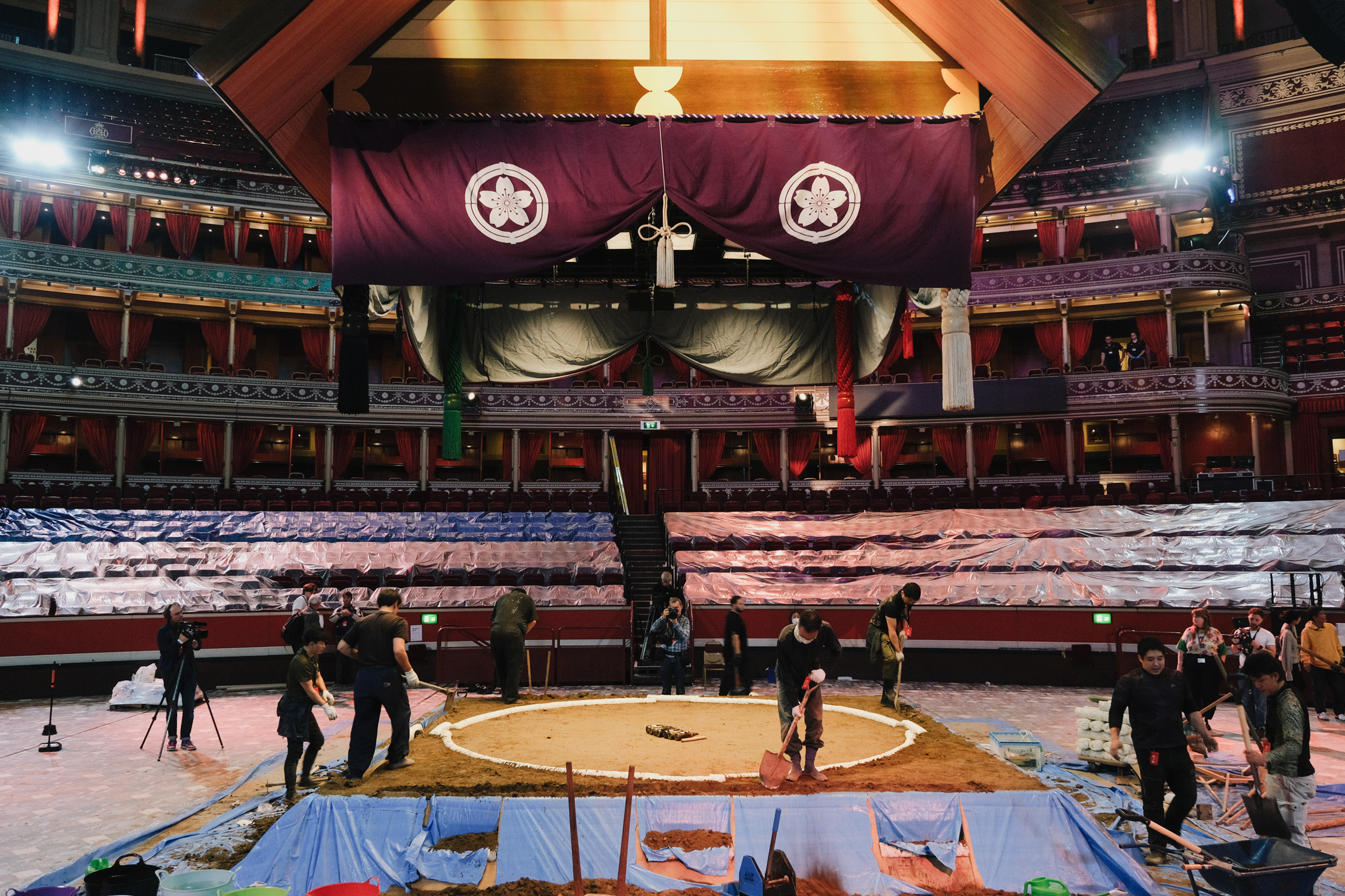 Interior of historic theatre with ornate red velvet seating, decorative ceiling medallions, and workers setting up central stage area.