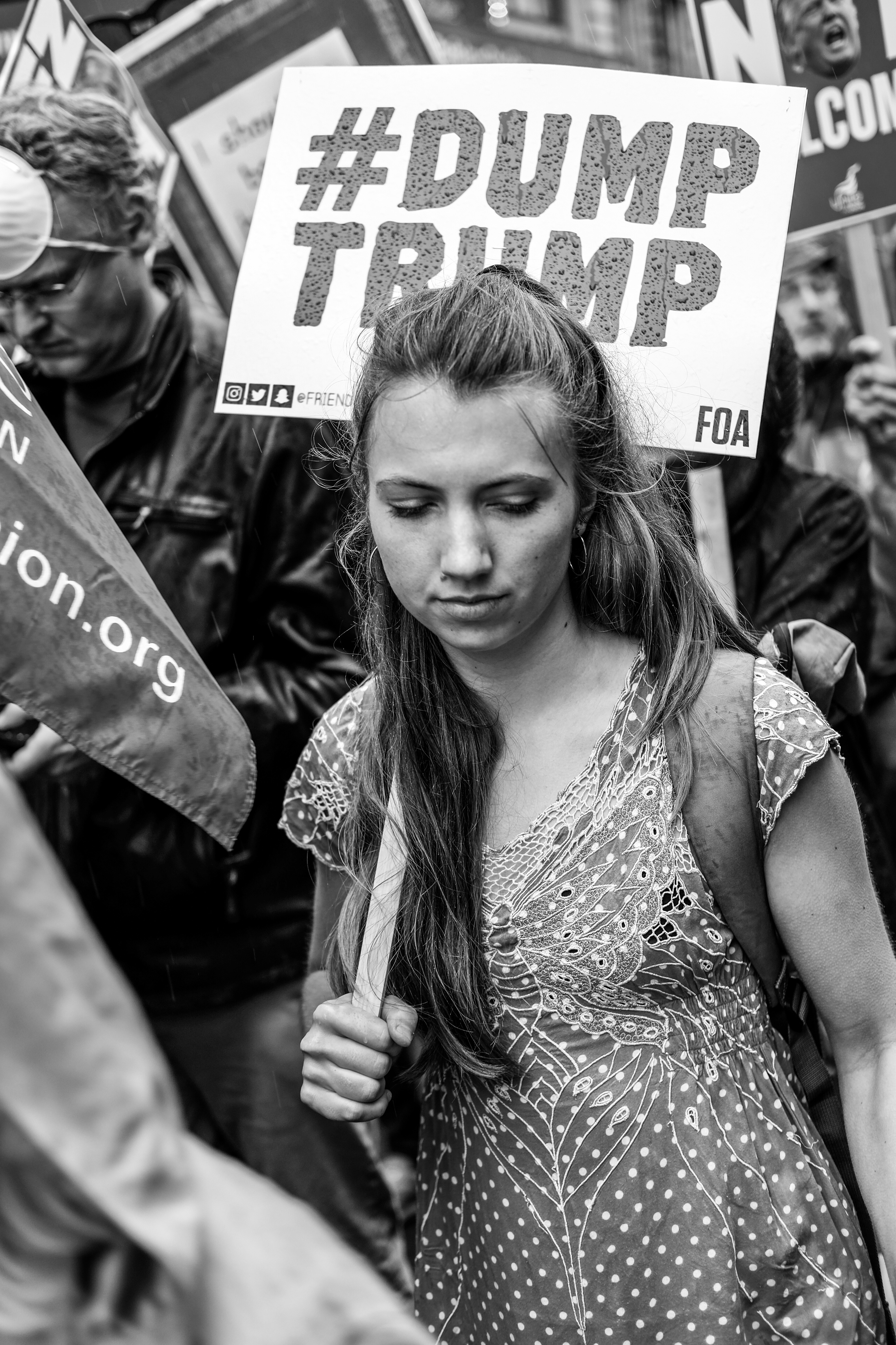 Black and white image showing a woman holding a #DUMP TRUMP sign at a protest rally. The woman has long hair and appears to be looking down thoughtfully.