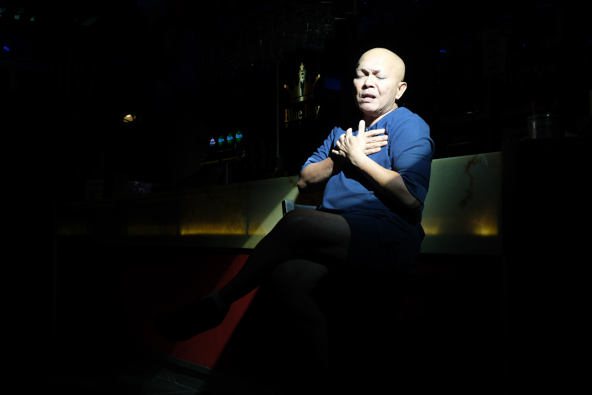Bald man in blue shirt sits on stage with hands on chest, illuminated by spotlight against dark background.