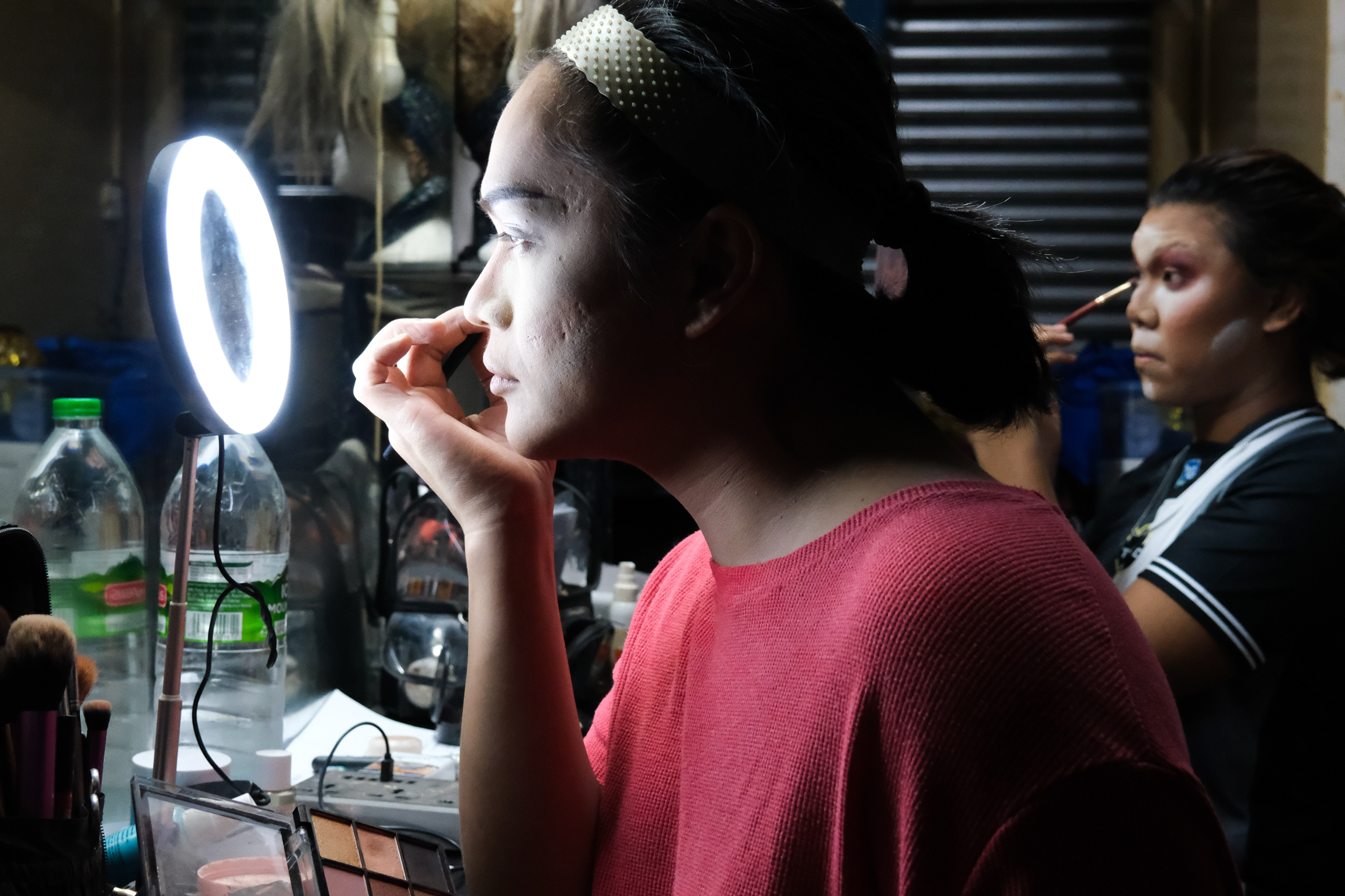 Woman in red top applying makeup with ring light, another person visible in background of cluttered indoor space.