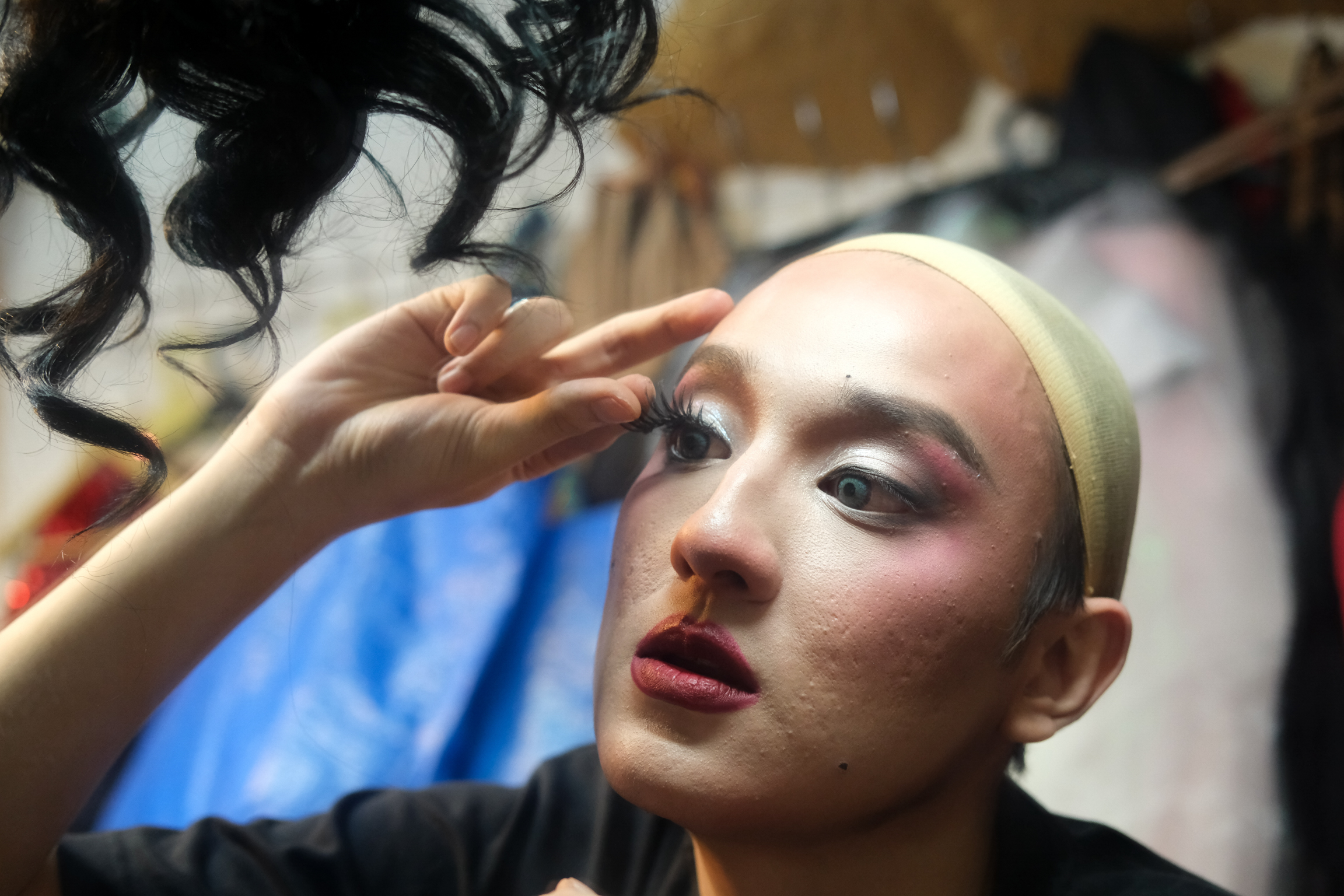 Person in bald cap applying stage makeup, hand positioned near eye area, colourful costumes visible in background.