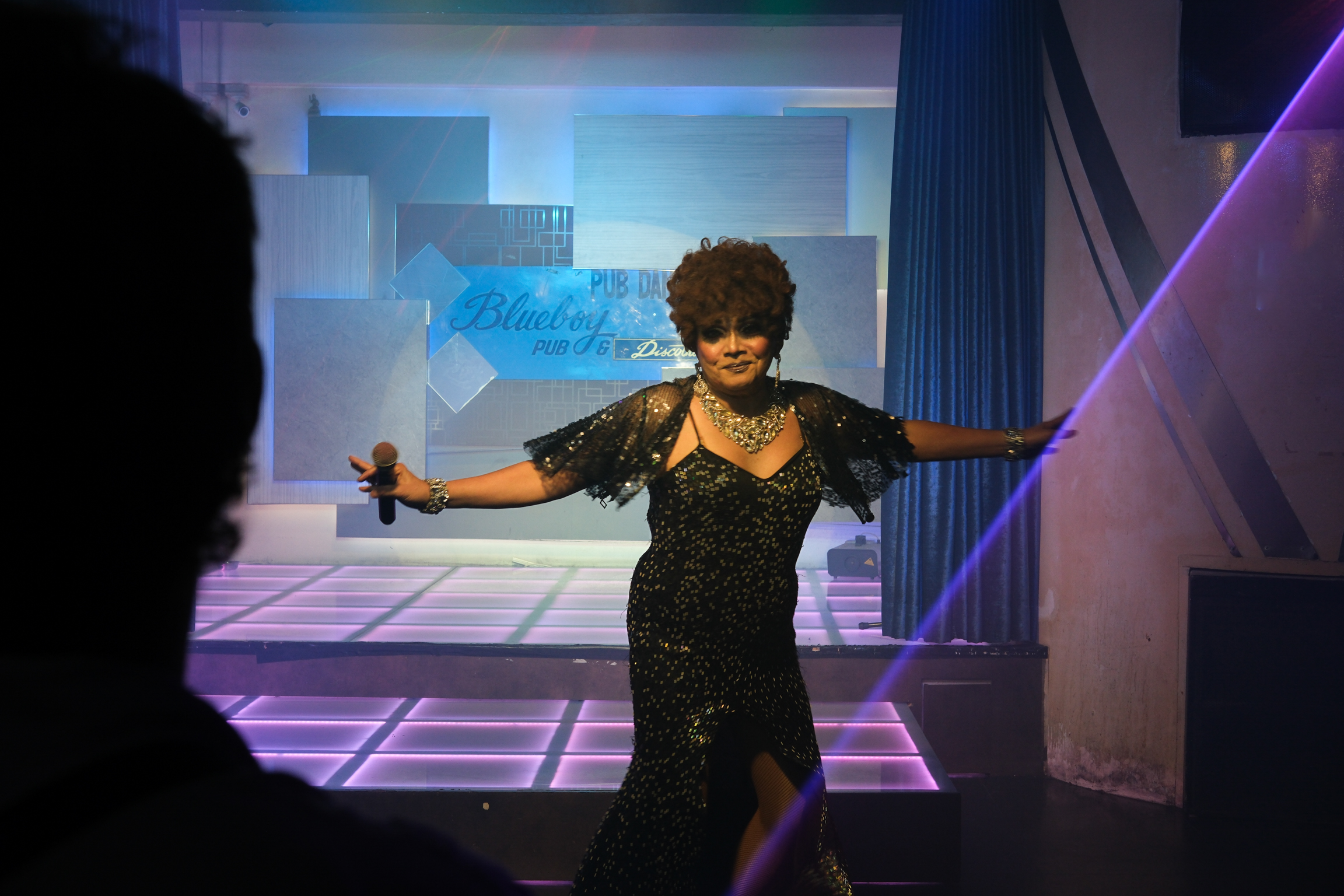 Performer in sequined black dress with arms outstretched on stage with purple lighting and geometric backdrop.