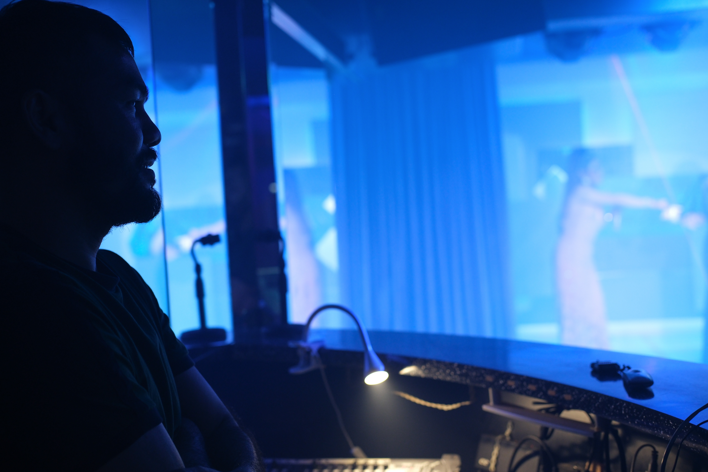 Person silhouetted in profile operating equipment with multiple blue-lit screens and monitors in darkened room.