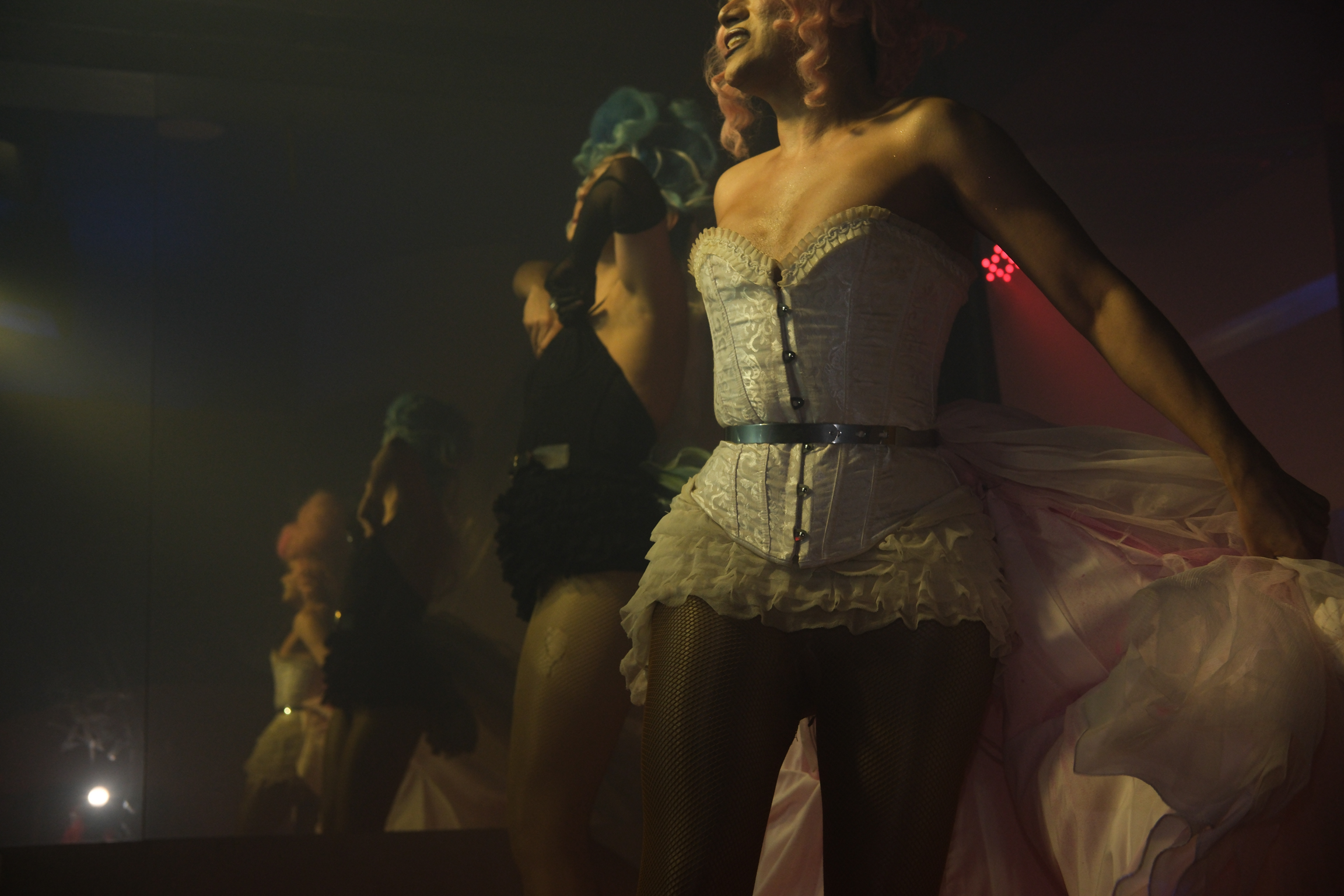 Performers in white corsets and tutus on dimly lit stage with warm golden lighting and dark background.
