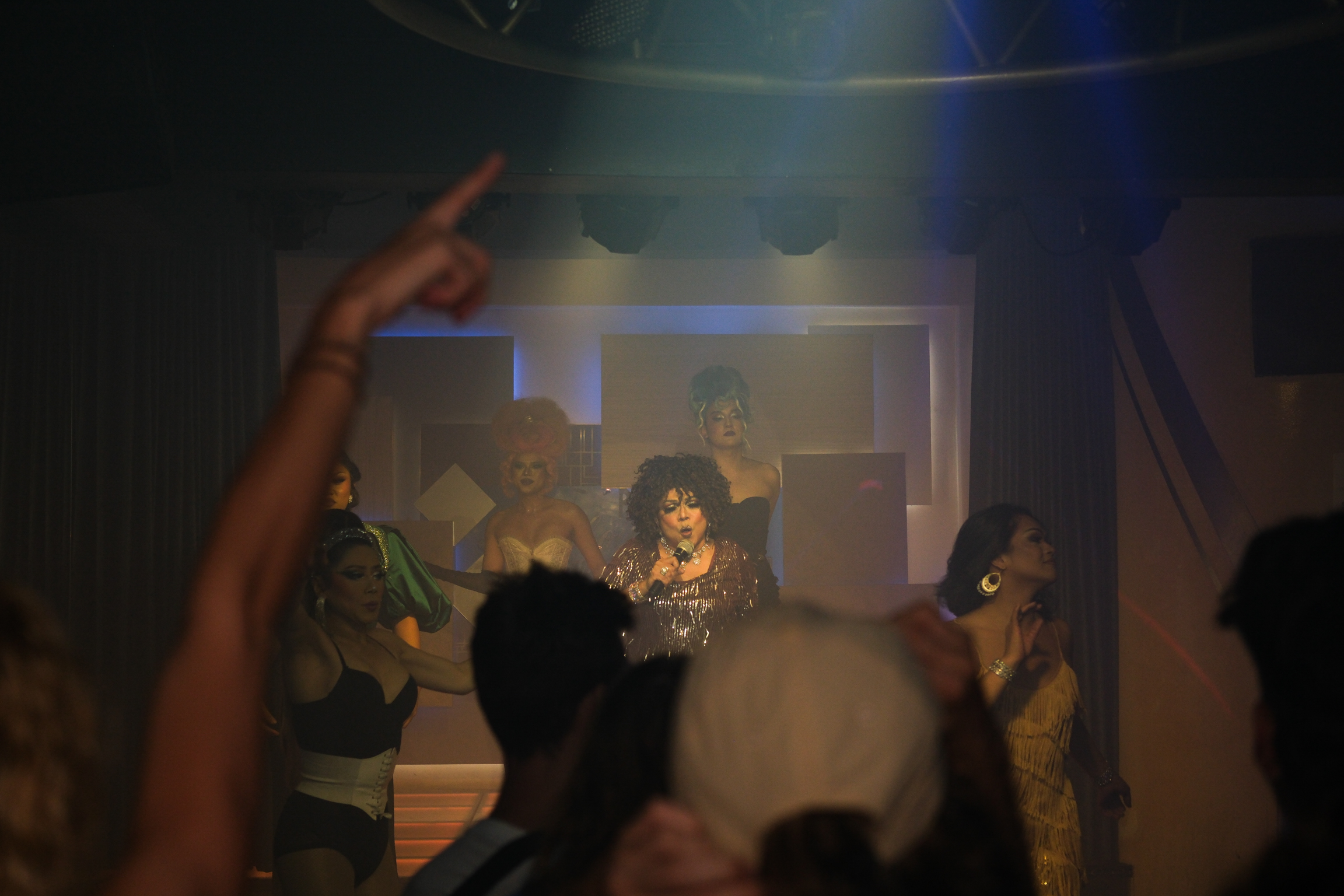 Dimly lit indoor scene with silhouetted figures in foreground, blue and purple stage lighting illuminating a performer in the background.