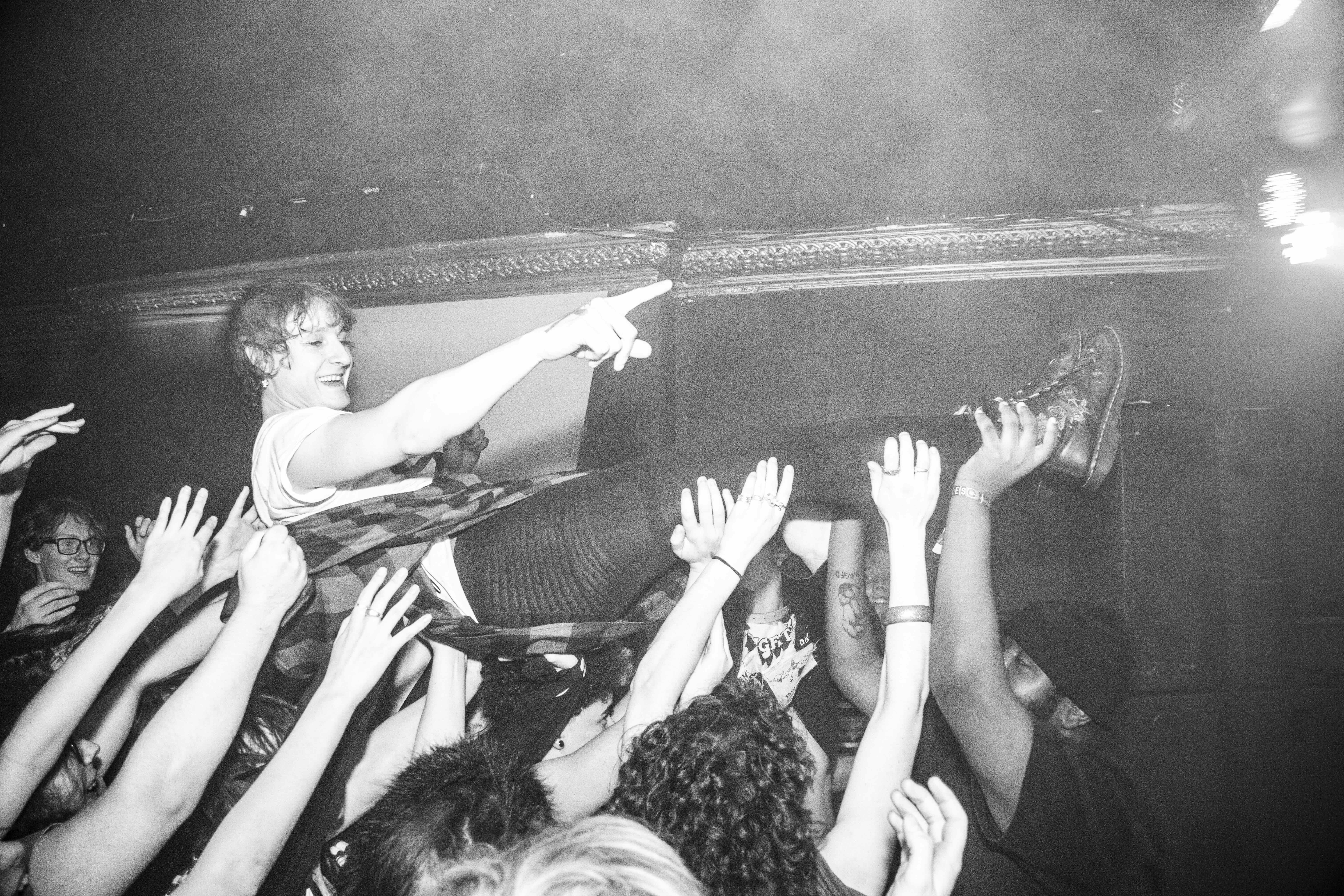 Black and white image of performer on stage pointing towards crowd with raised hands reaching upwards, smoky atmosphere visible.