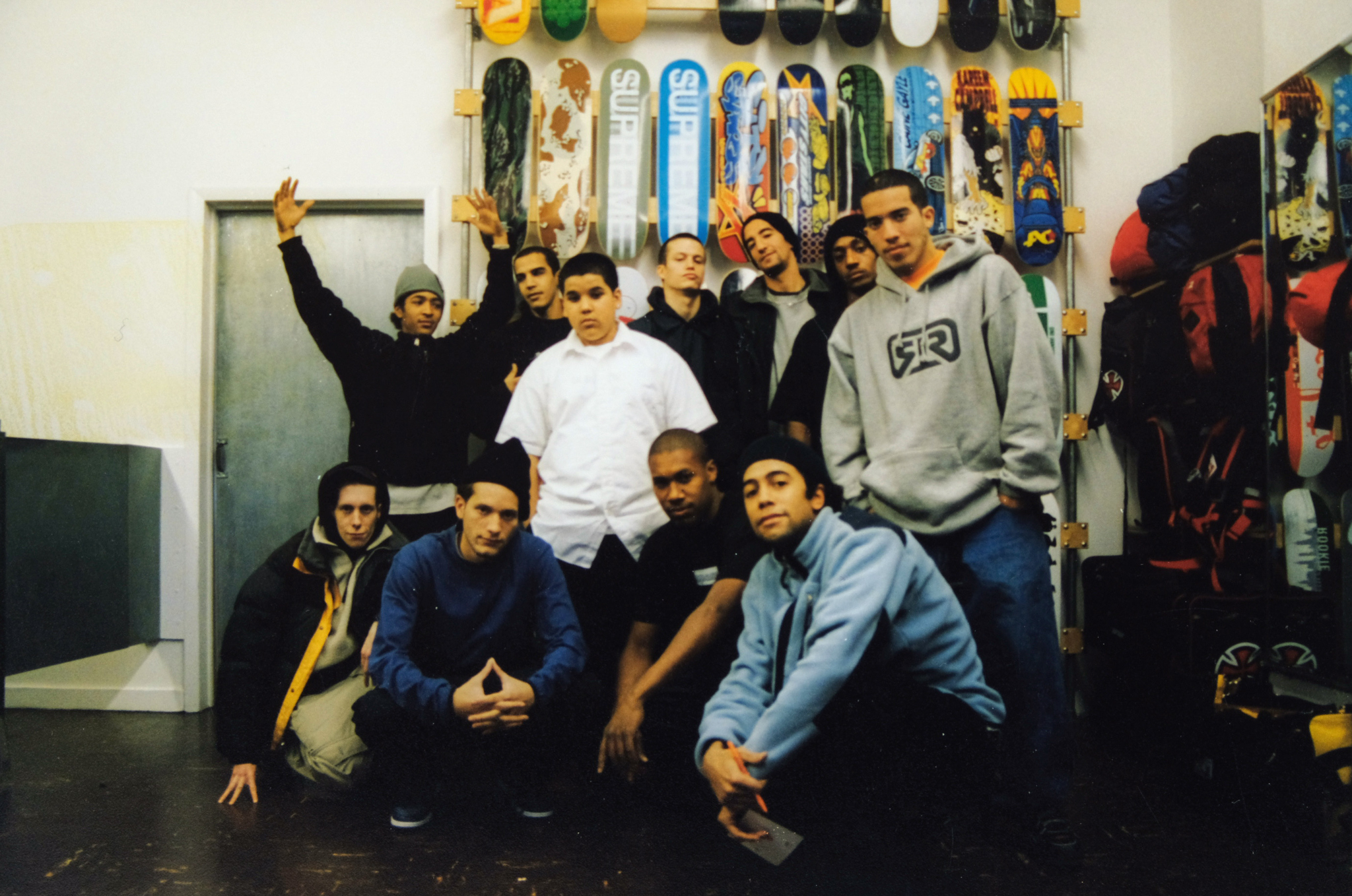 Group of young men posing in skate shop with skateboard decks mounted on white wall behind them.