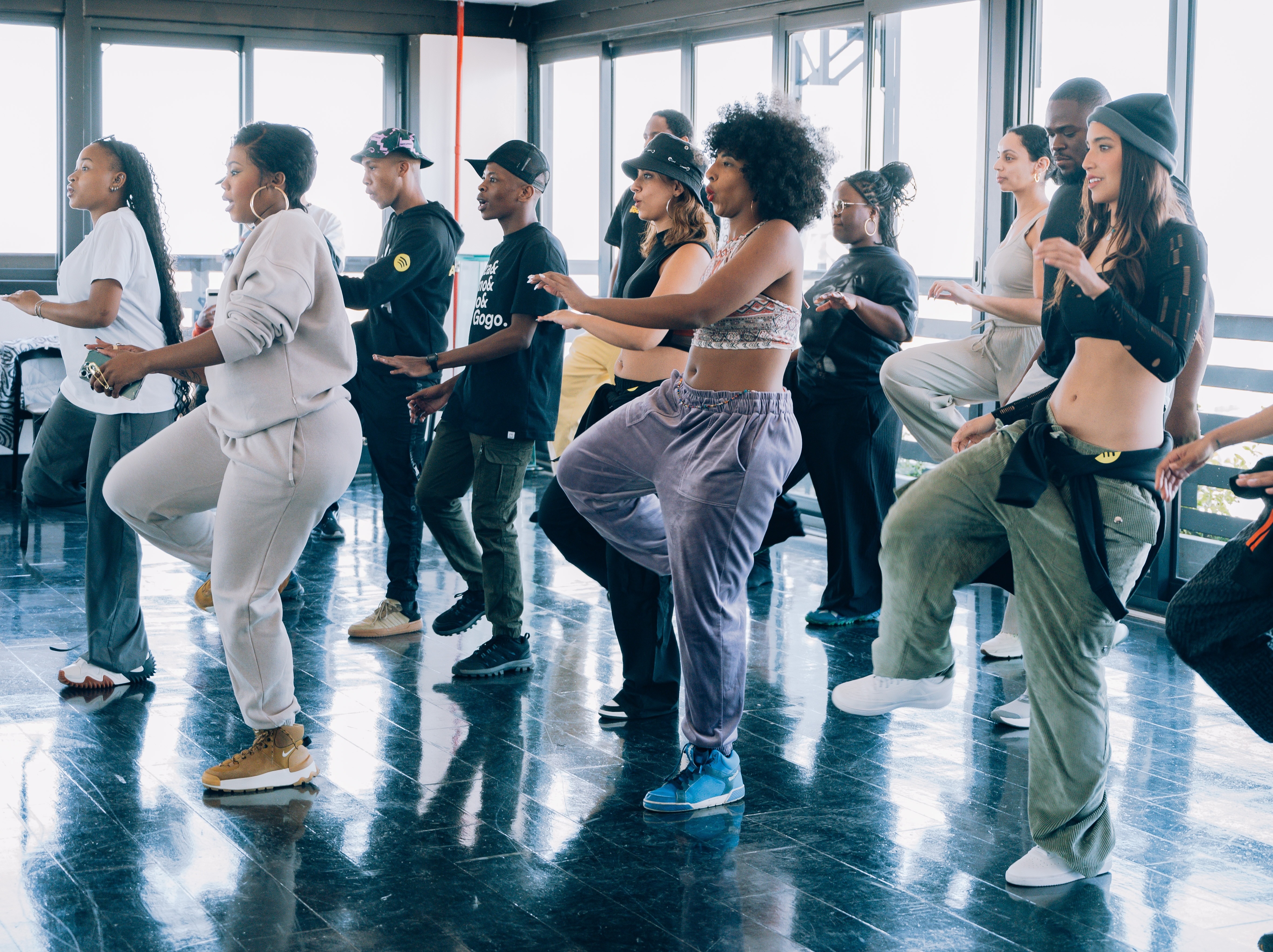 Group of people dancing in a studio, wearing casual outfits in various colours, some shirtless, against a large window background.