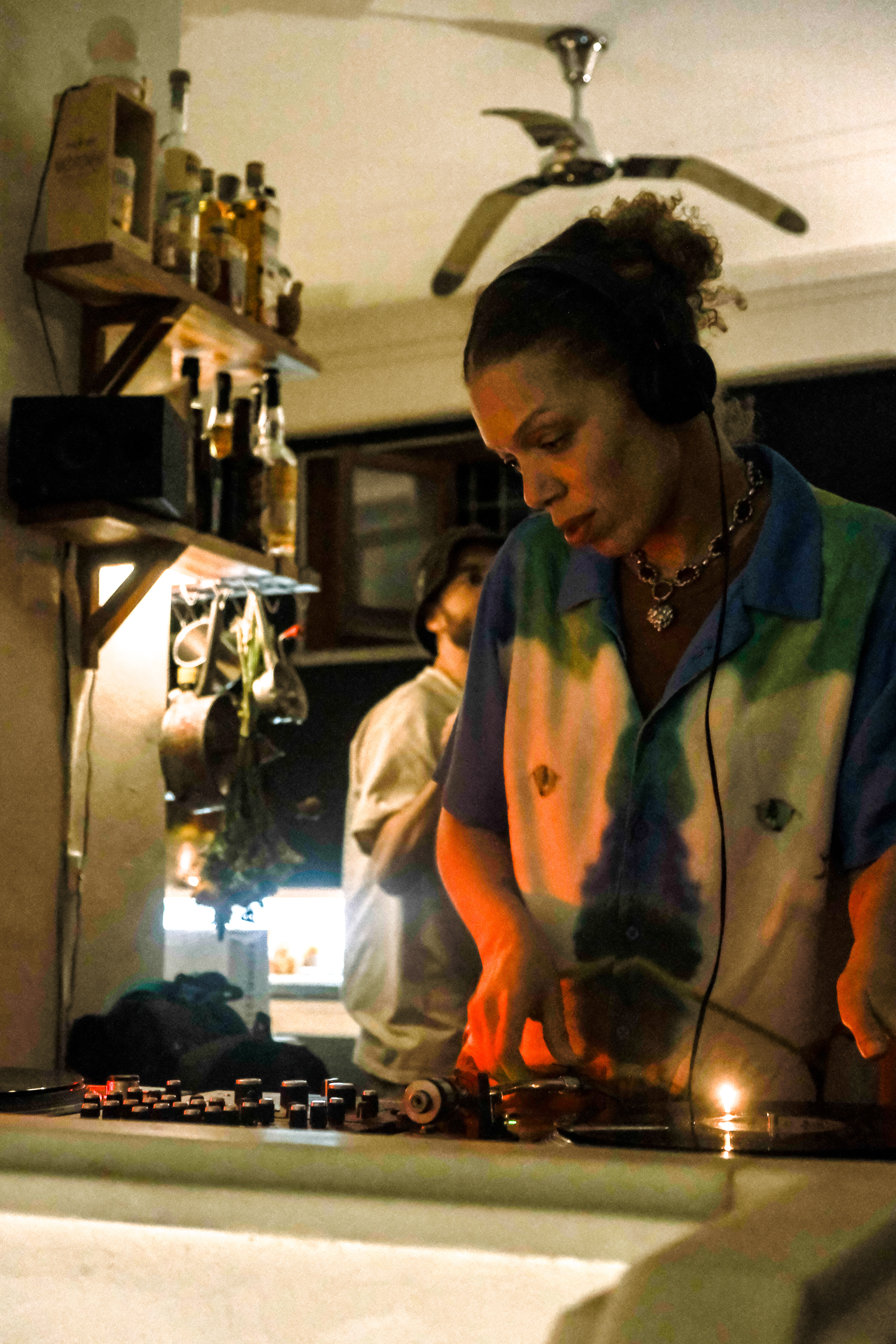 Woman in colourful shirt DJing at kitchen counter with mixing equipment, warm orange lighting, bottles on shelves above