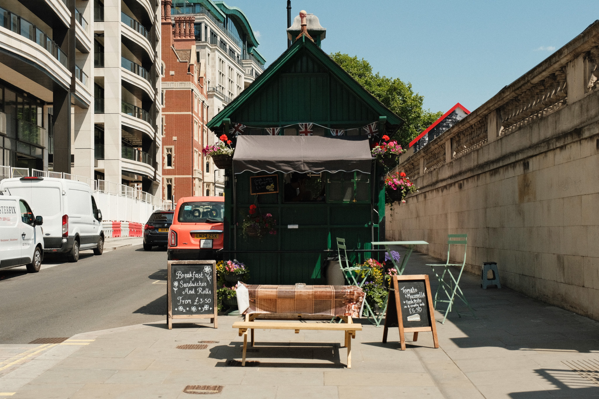 Green market stall with striped awning on urban street, flanked by chalkboard signs and wooden benches, surrounded by mixed architecture.