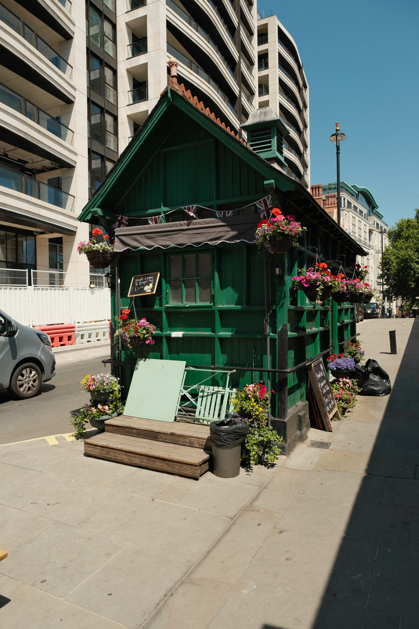 Green Victorian-style beach hut with decorative trim and flower boxes on urban street, modern apartment buildings behind.