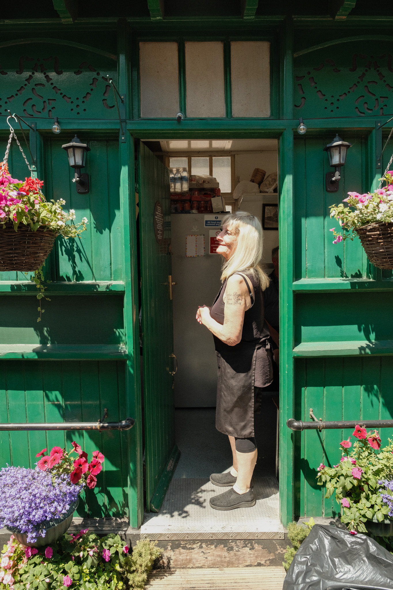 Woman with blonde hair in black top and grey apron standing in doorway of bright green painted shop with hanging flower baskets.