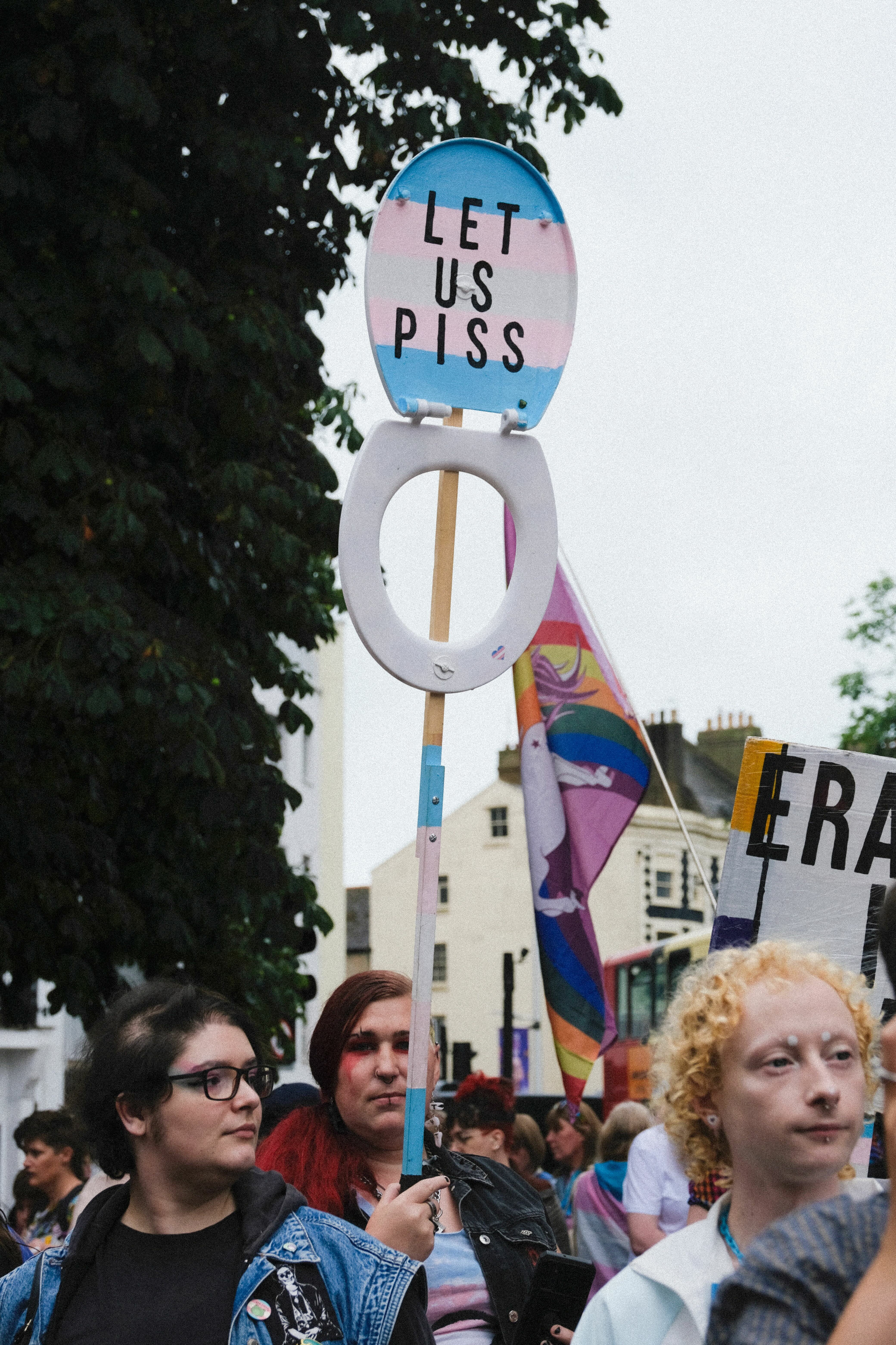 Protest crowd with person holding circular blue and pink sign reading "LET US PISS" beneath overcast sky and leafy trees.