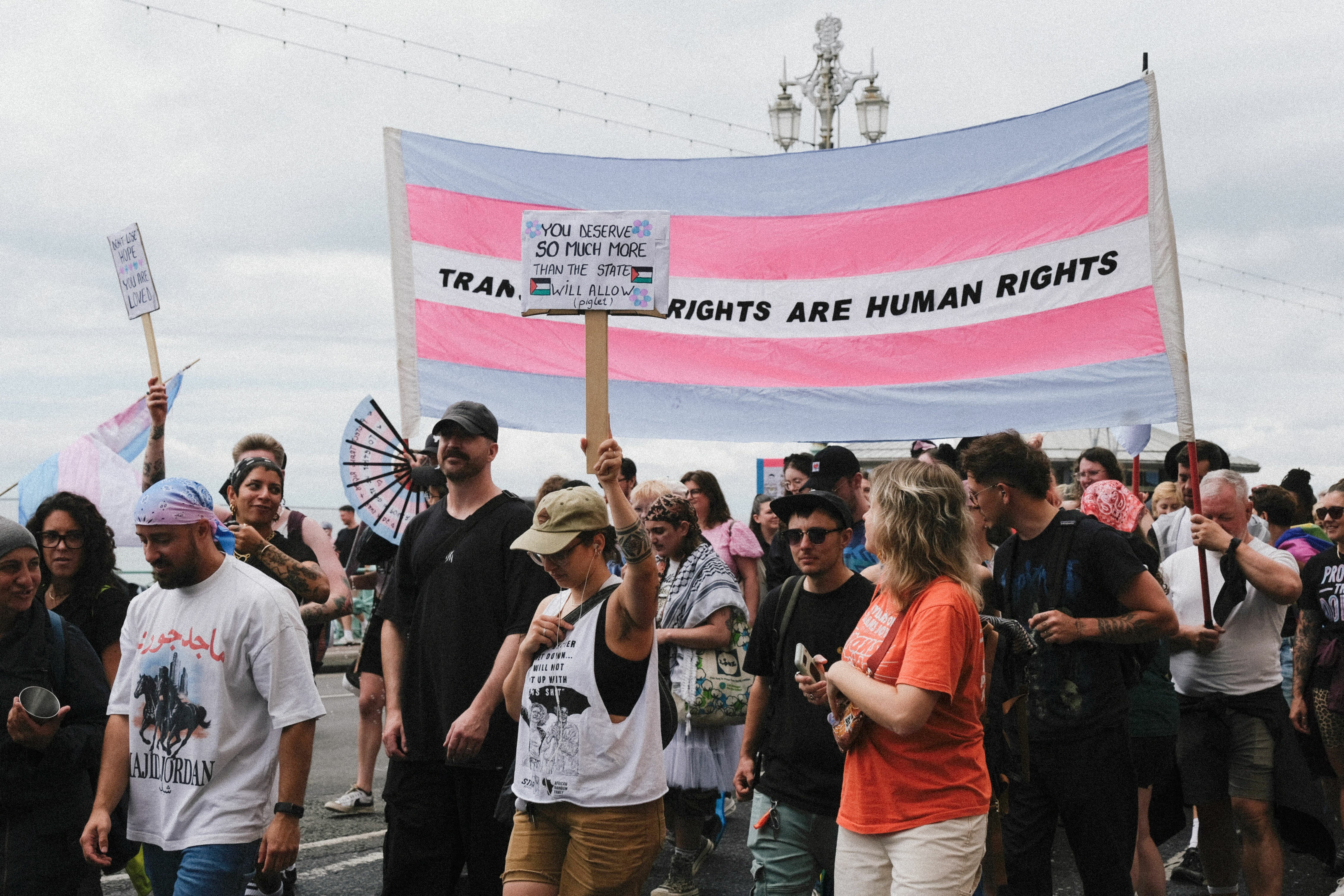 Crowd of protesters holding large transgender flag banner reading "TRANS RIGHTS ARE HUMAN RIGHTS" with people in casual clothing beneath.