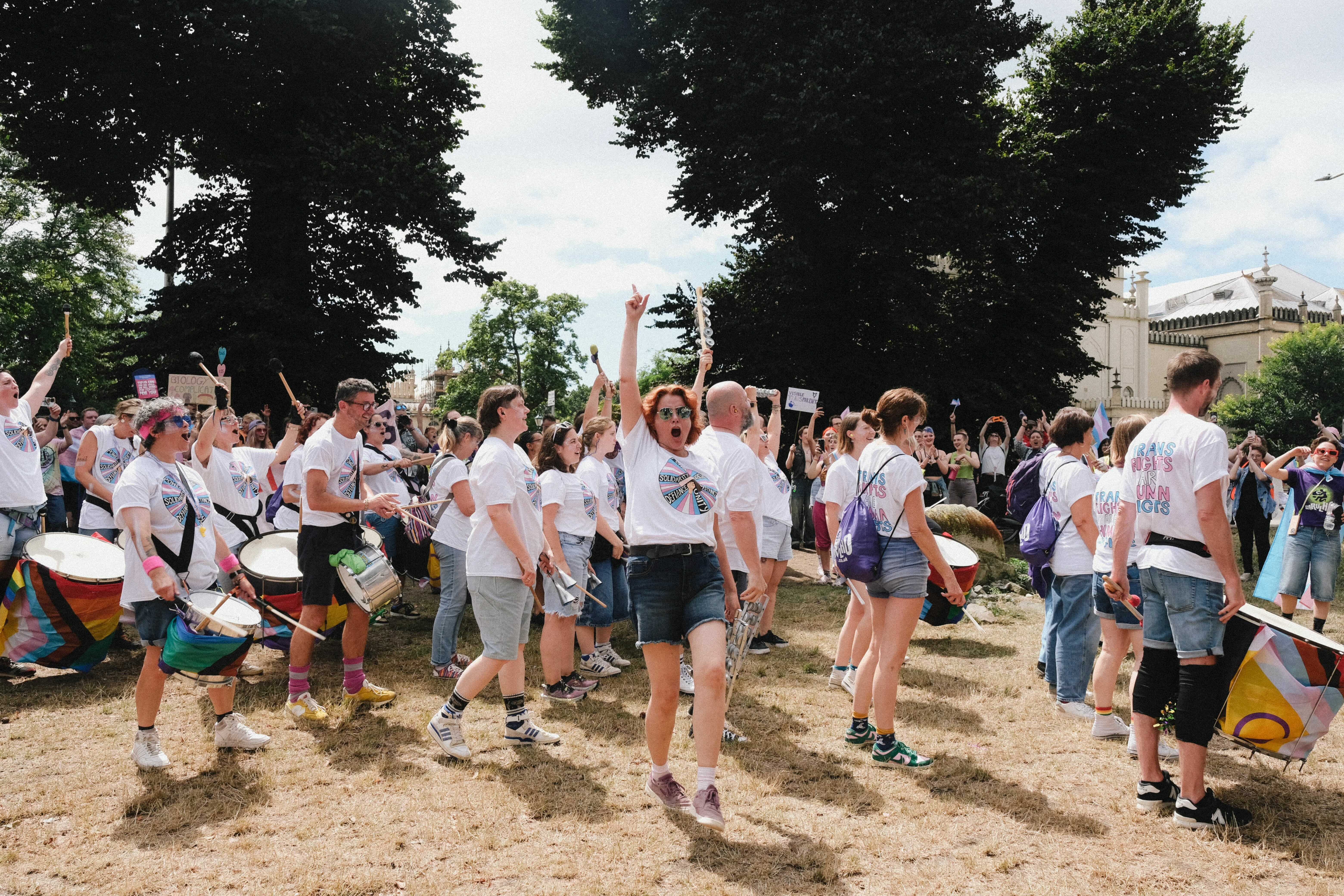 Large group of children in white t-shirts holding drumsticks and drums outdoors on dirt ground, surrounded by trees and buildings.