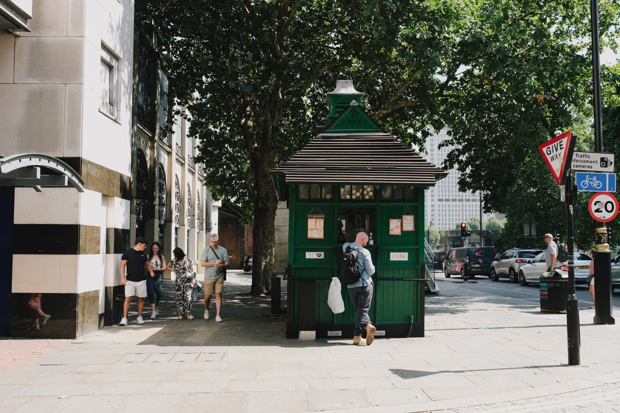 Green Victorian police box on urban pavement with people nearby, surrounded by trees and buildings with road signs visible.