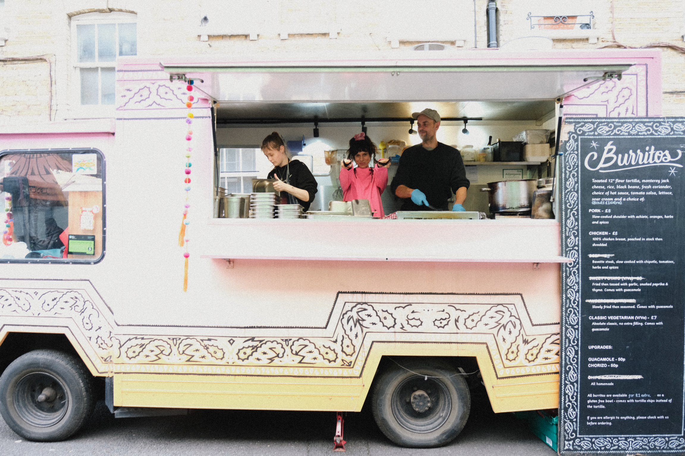 Pink and cream food truck with ornate white decorative patterns. Three people visible in serving window. Black menu board on right side.