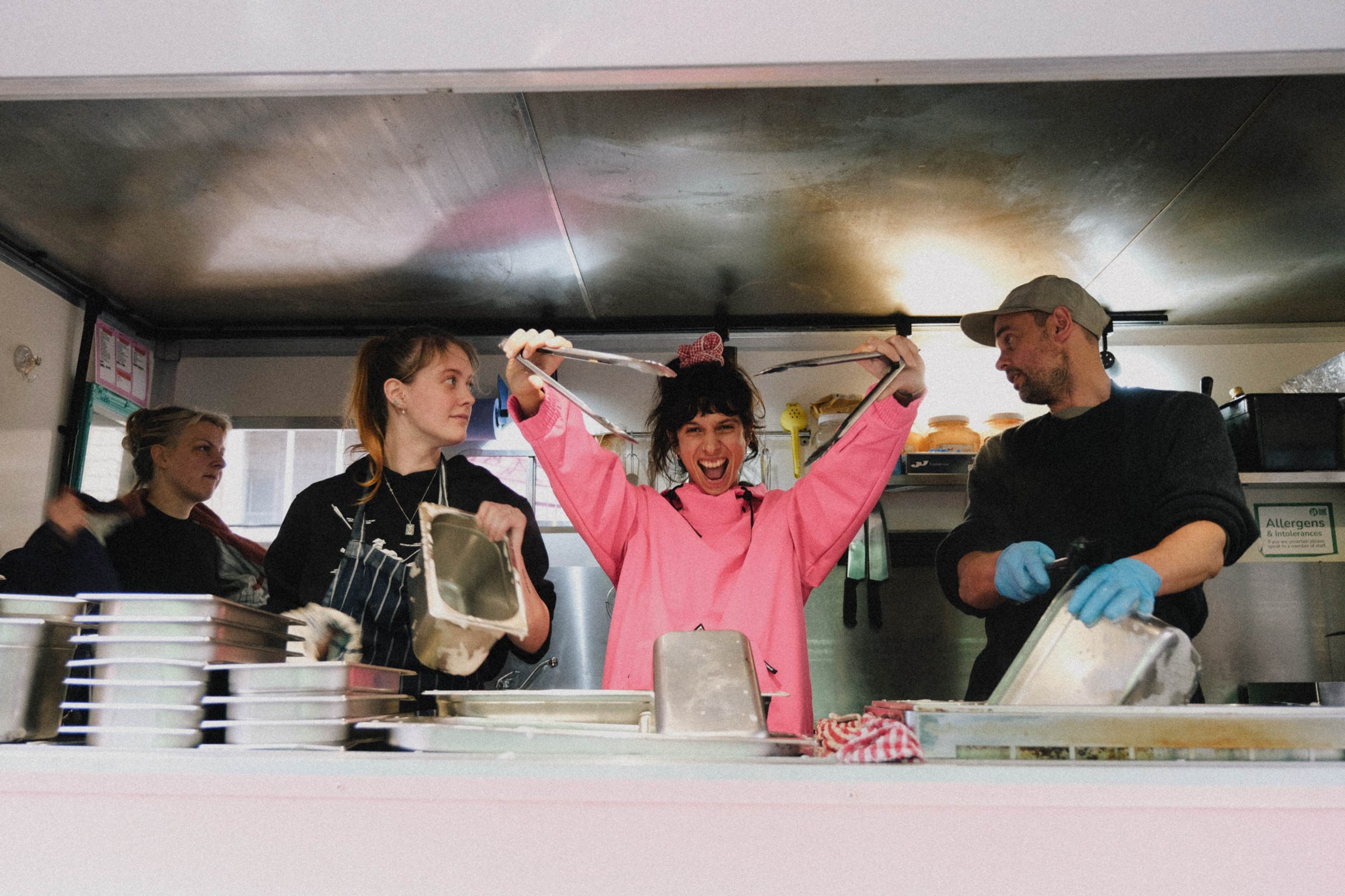 Three people working in food truck kitchen, person in centre wearing bright pink hoodie with arms raised, flanked by two others in dark clothing.
