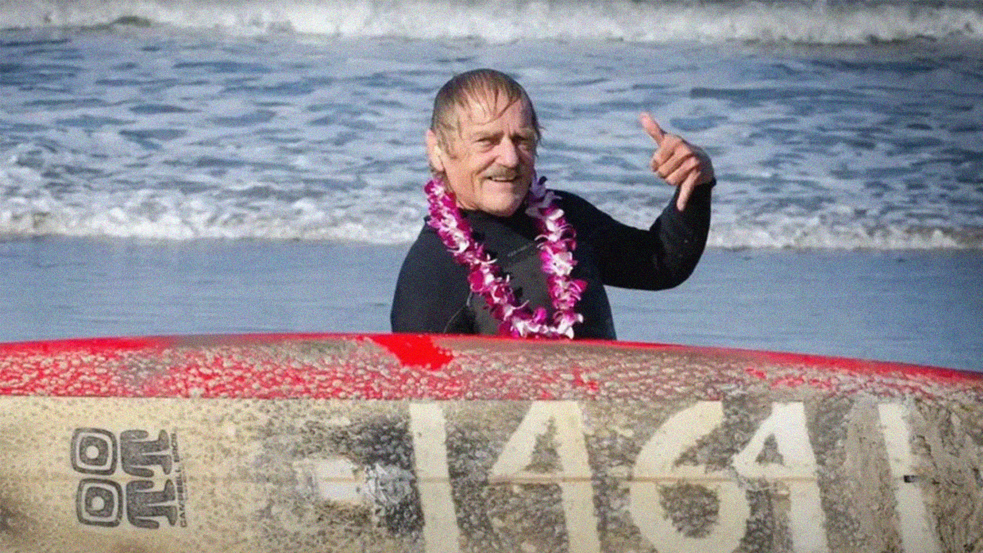 Man in wetsuit and pink lei giving thumbs up behind large rock marked "1641" with red ribbon on beach