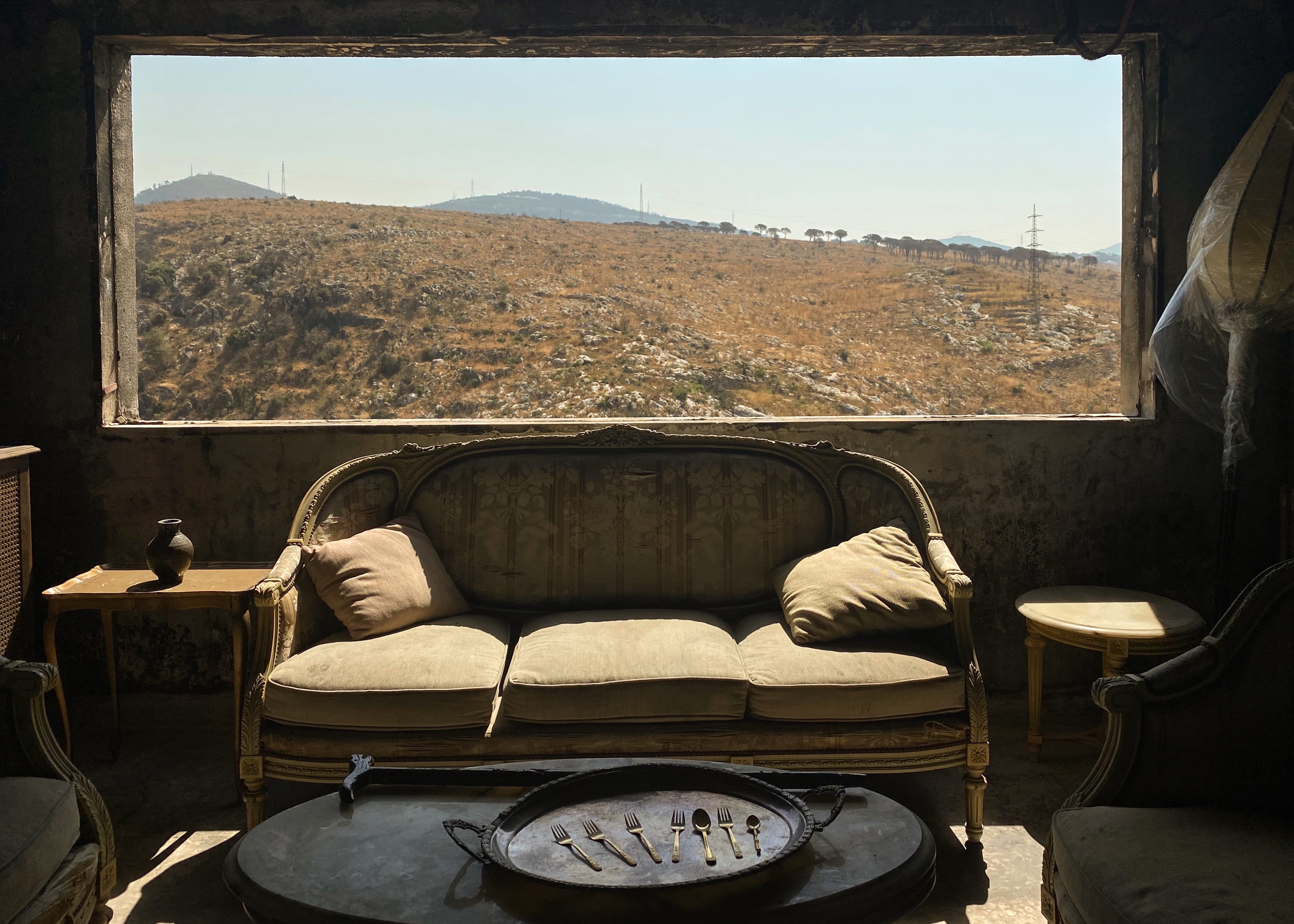 Wide panoramic view of a desert landscape with hills and mountains through a window frame, with a dark vintage sofa in the foreground.