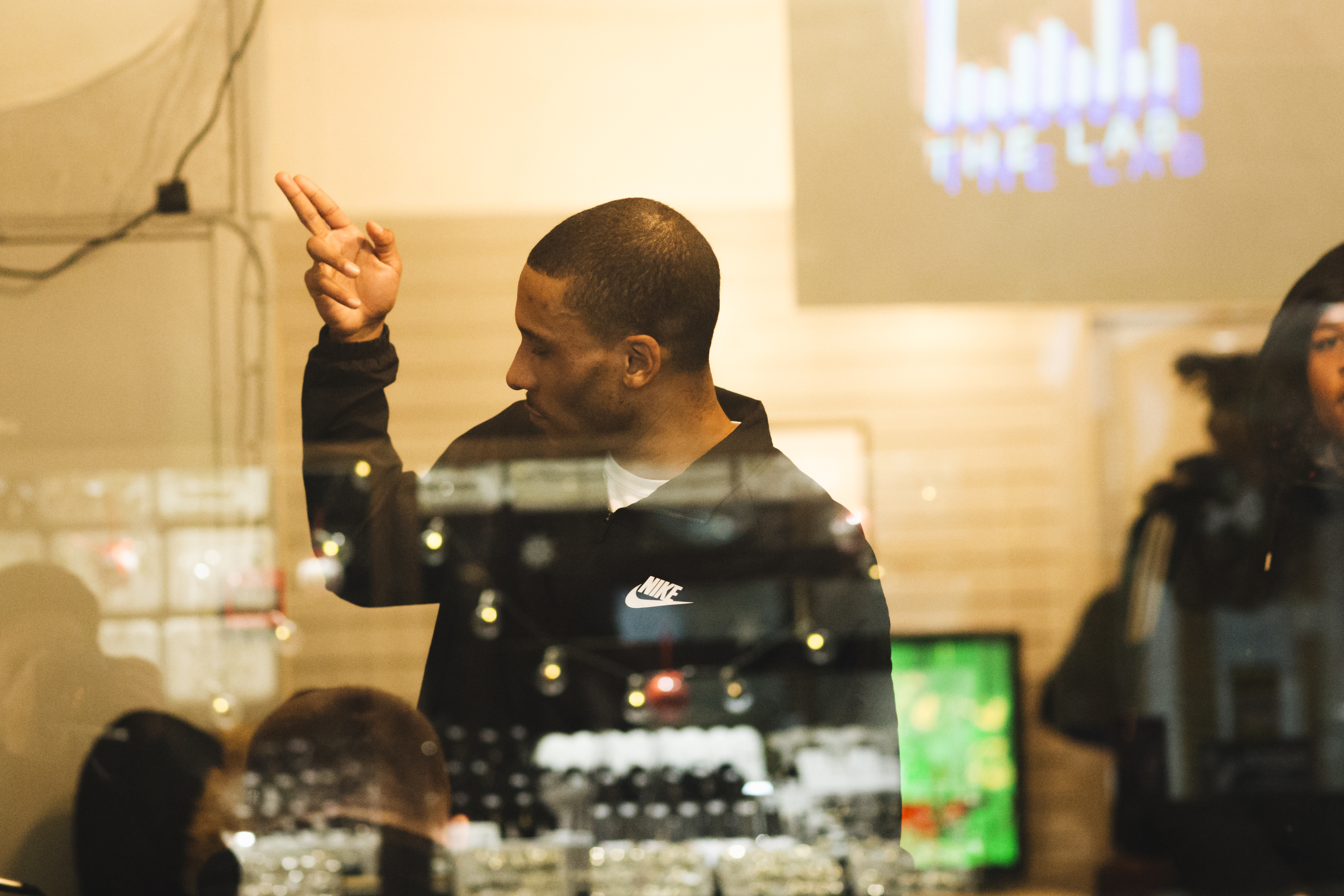 Man in dark jacket pointing upward behind glass with reflections and lights, blurred figures in background