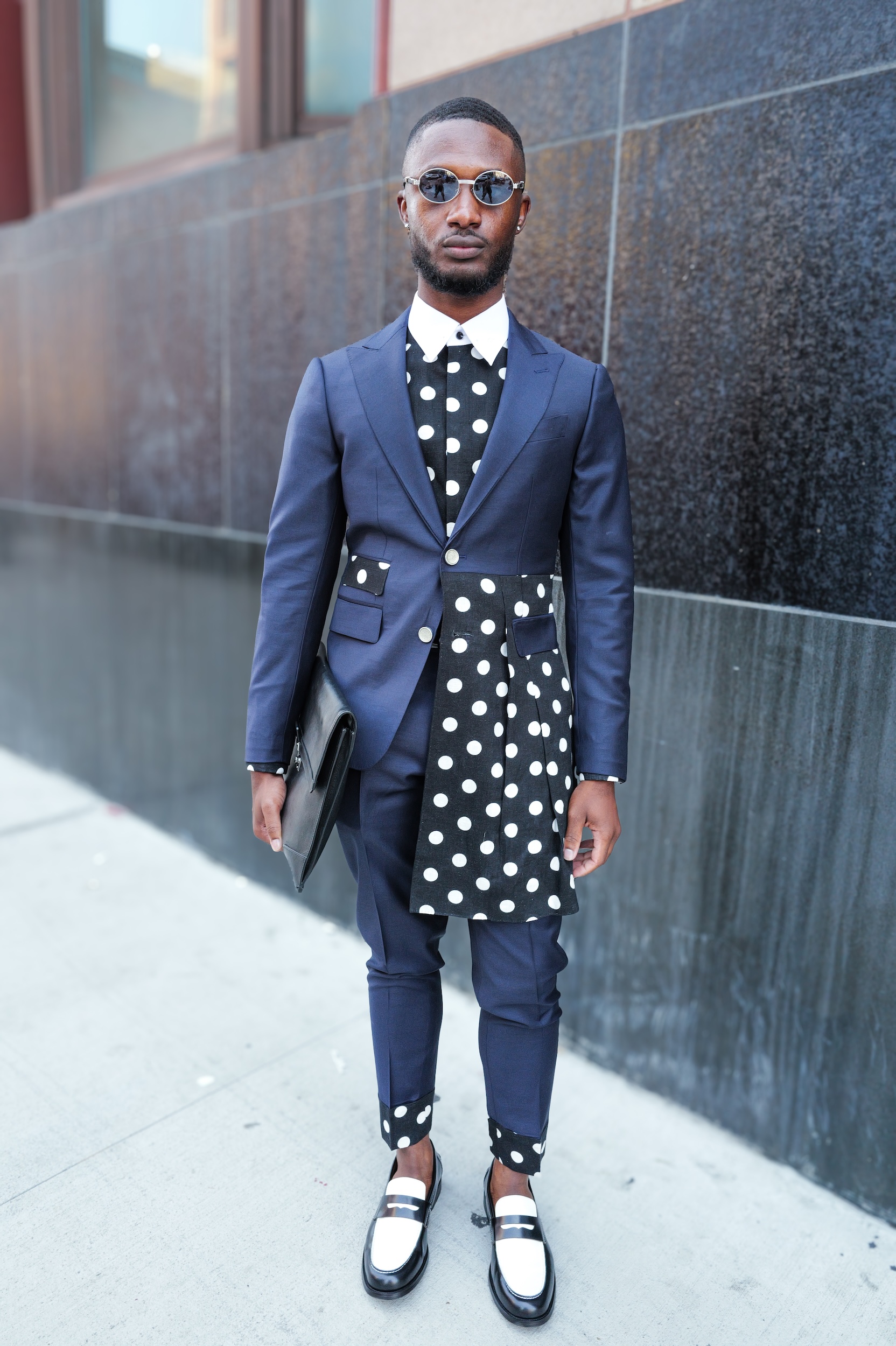 Man in navy suit with black polka dot waistcoat and bow tie, wearing sunglasses and black-white Oxford shoes on pavement.