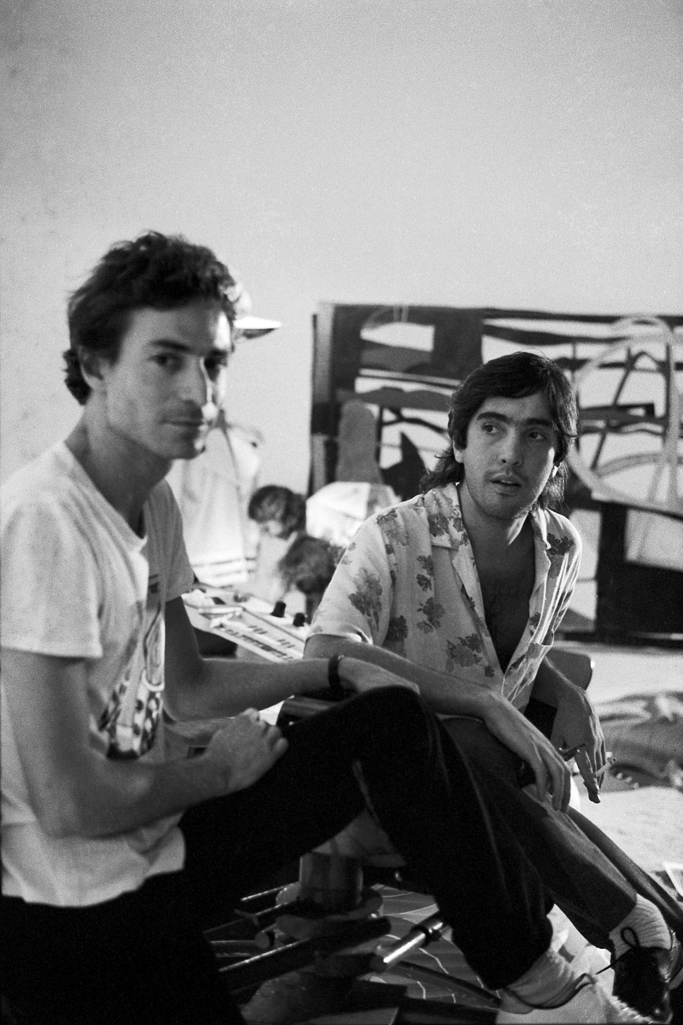 Two young men sitting on wooden chairs, wearing casual clothing. Black and white photograph with a slightly gloomy, pensive atmosphere.