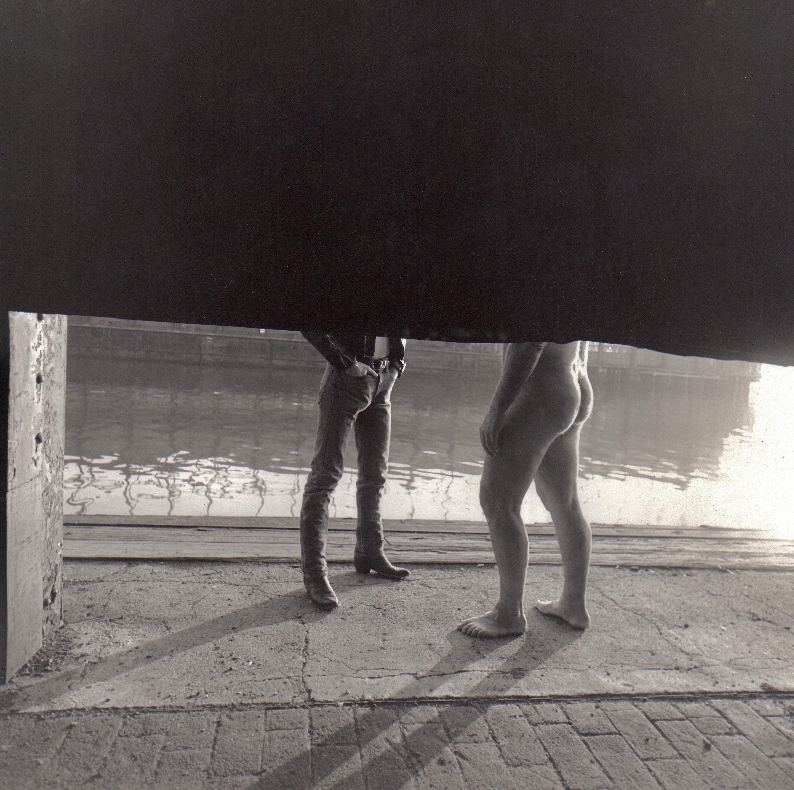 Two pairs of legs beneath dark umbrella on wet cobblestone pavement, with white concrete barrier and water visible in background.