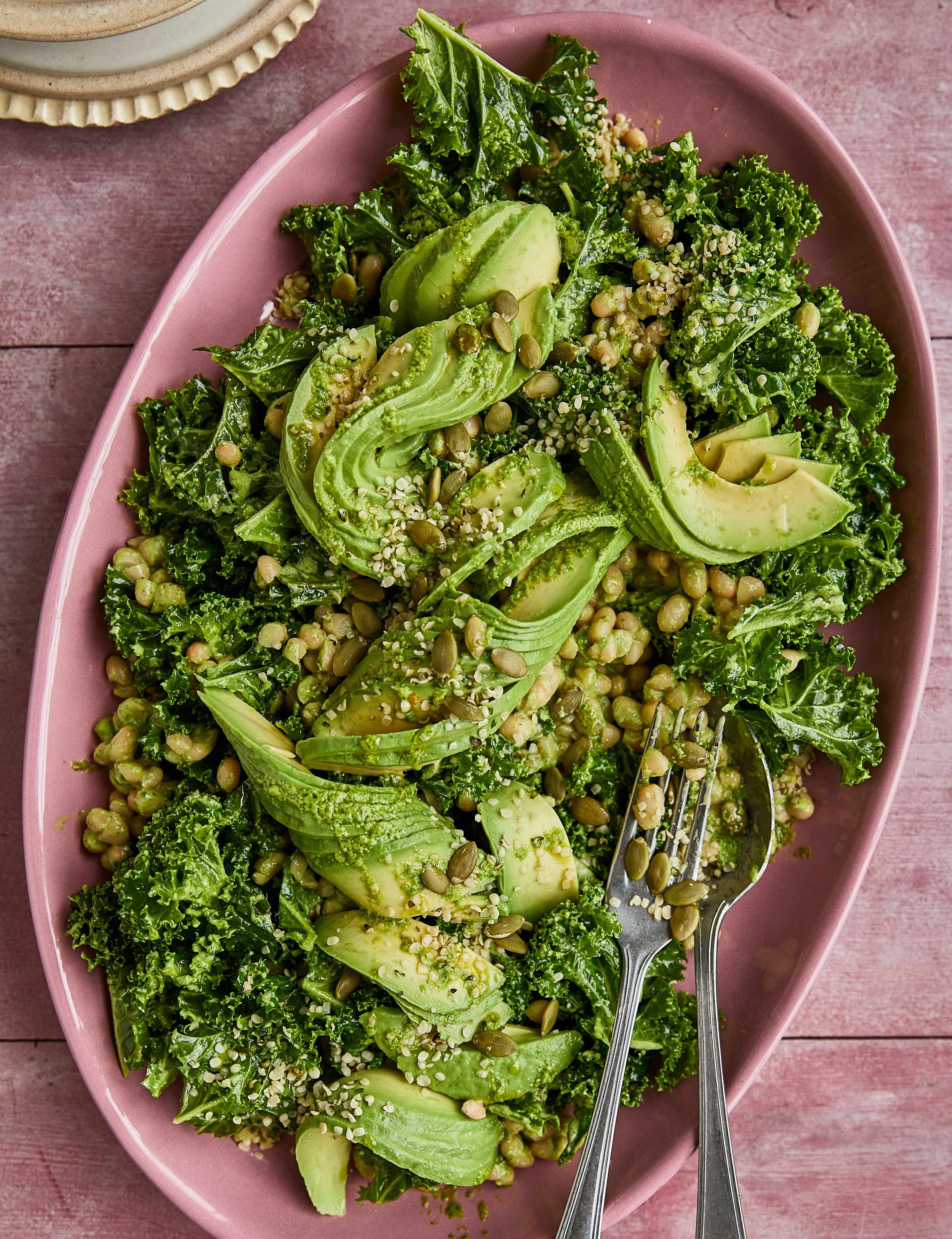 Pink oval bowl containing green kale salad with sliced avocado, small legumes, and white cheese crumbles, served with gold fork on wooden surface.