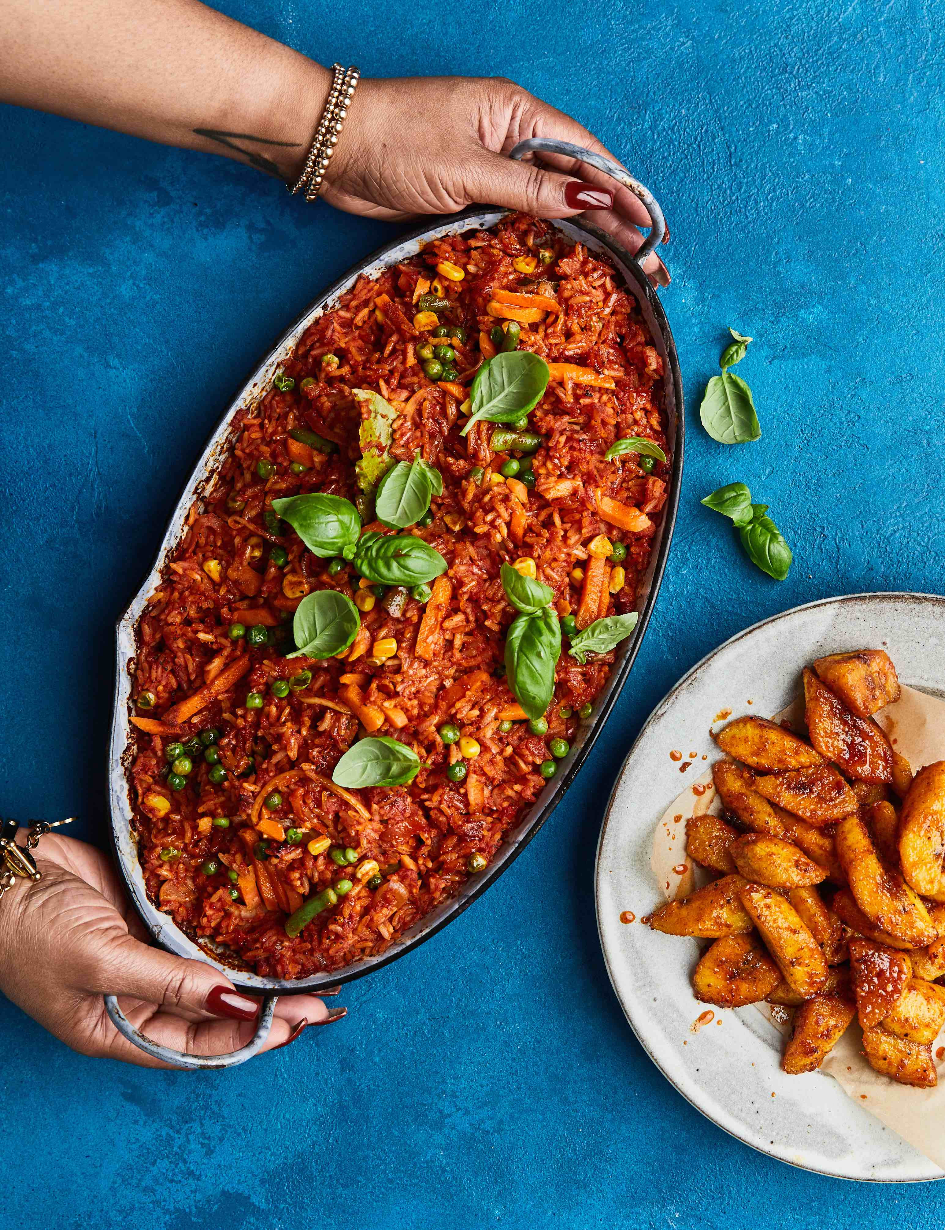 Hands holding oval baking dish with red tomato-based casserole topped with green basil leaves, served with roasted potato wedges on blue surface.