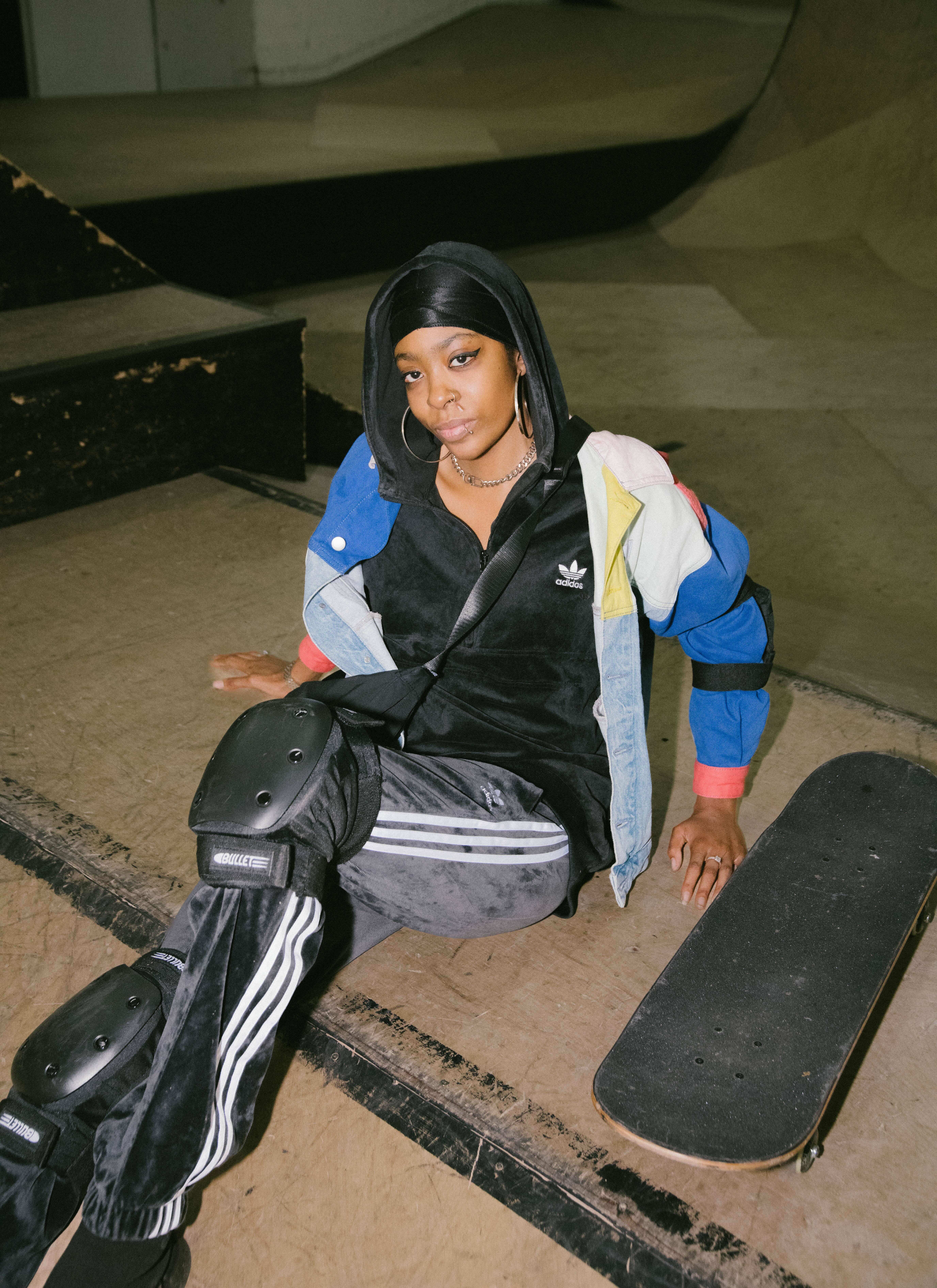Woman sitting on stairs holding a skateboard, wearing a colourful Adidas tracksuit with a hood.