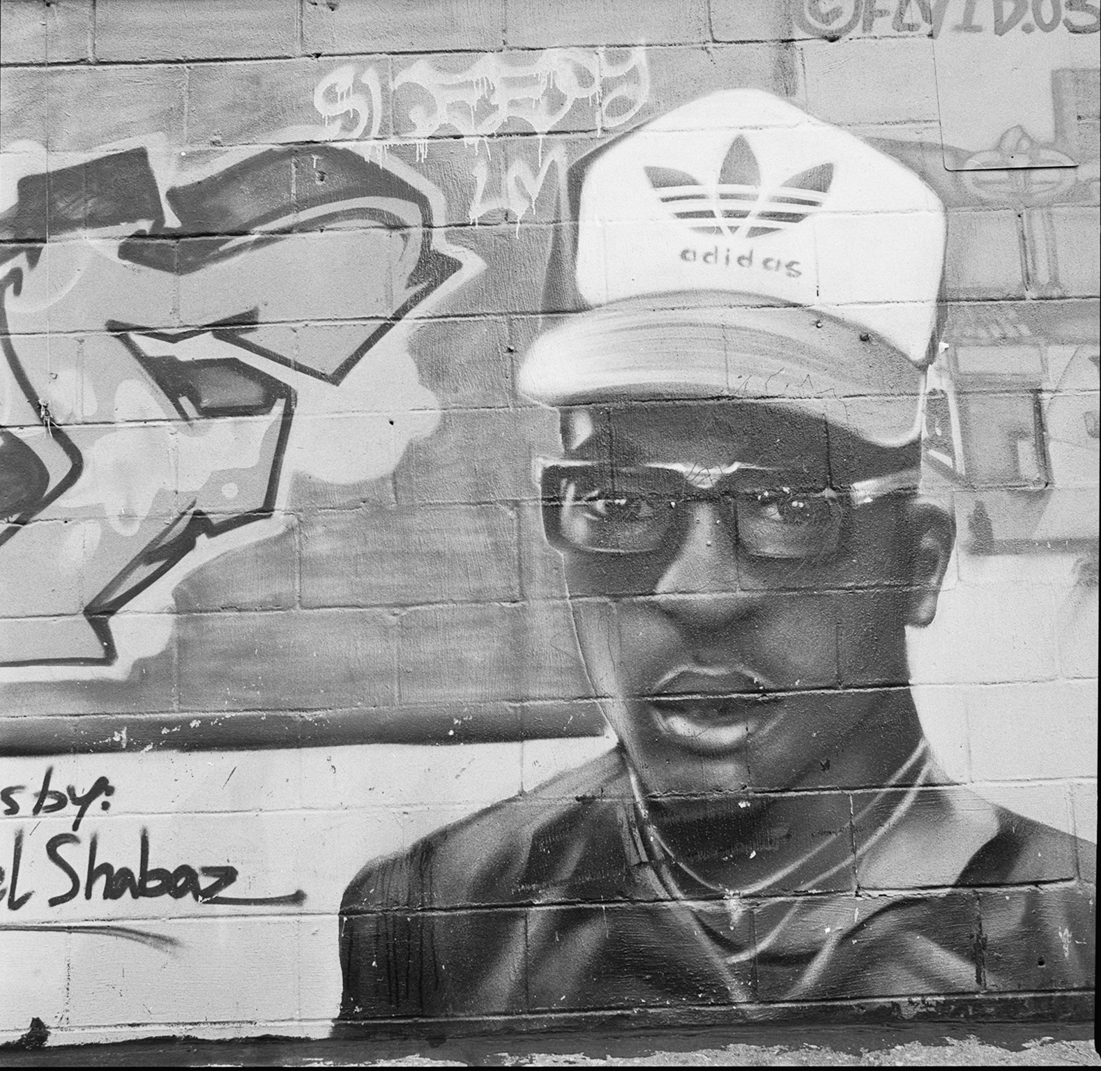 Black and white mural on brick wall showing man wearing glasses and Adidas cap, with graffiti-style text and abstract shapes.