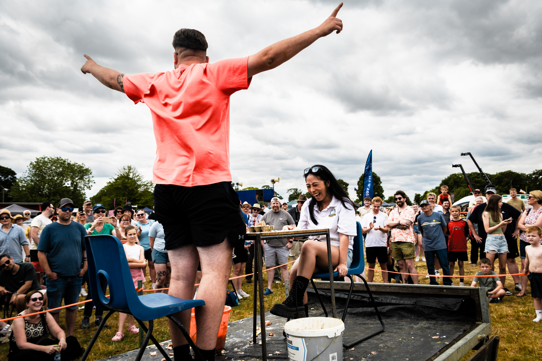 Man in orange shirt with arms raised standing over water tank, crowd watching behind barriers under cloudy sky.