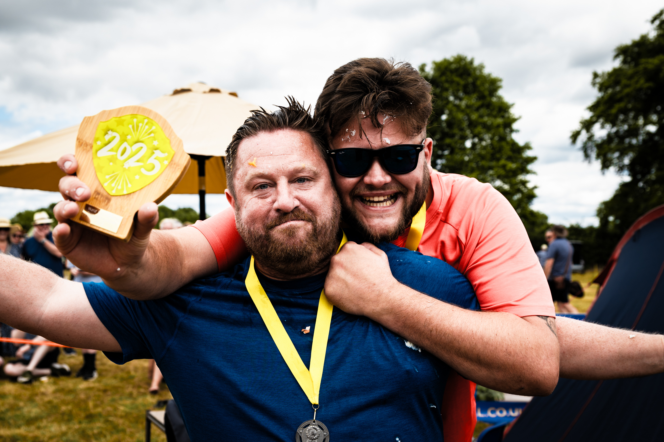 Two men embracing outdoors, one holding yellow medal displaying "2023", both wearing blue shirts, sunglasses, with crowd and trees behind.