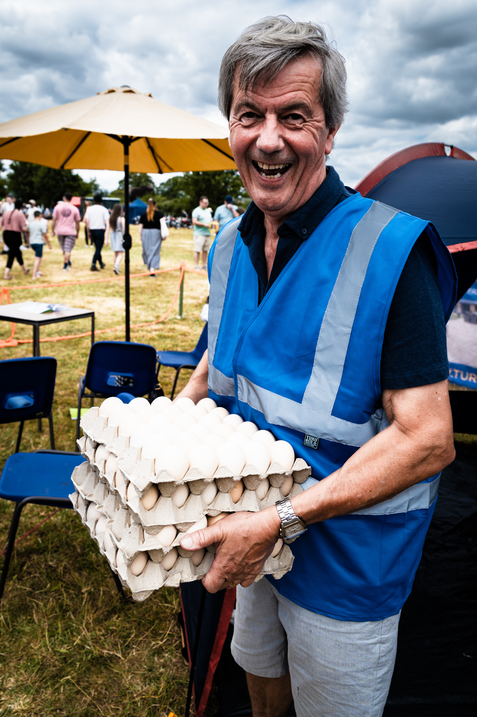 Man in blue high-vis vest holding stacked egg boxes at outdoor event with yellow parasol and people in background.