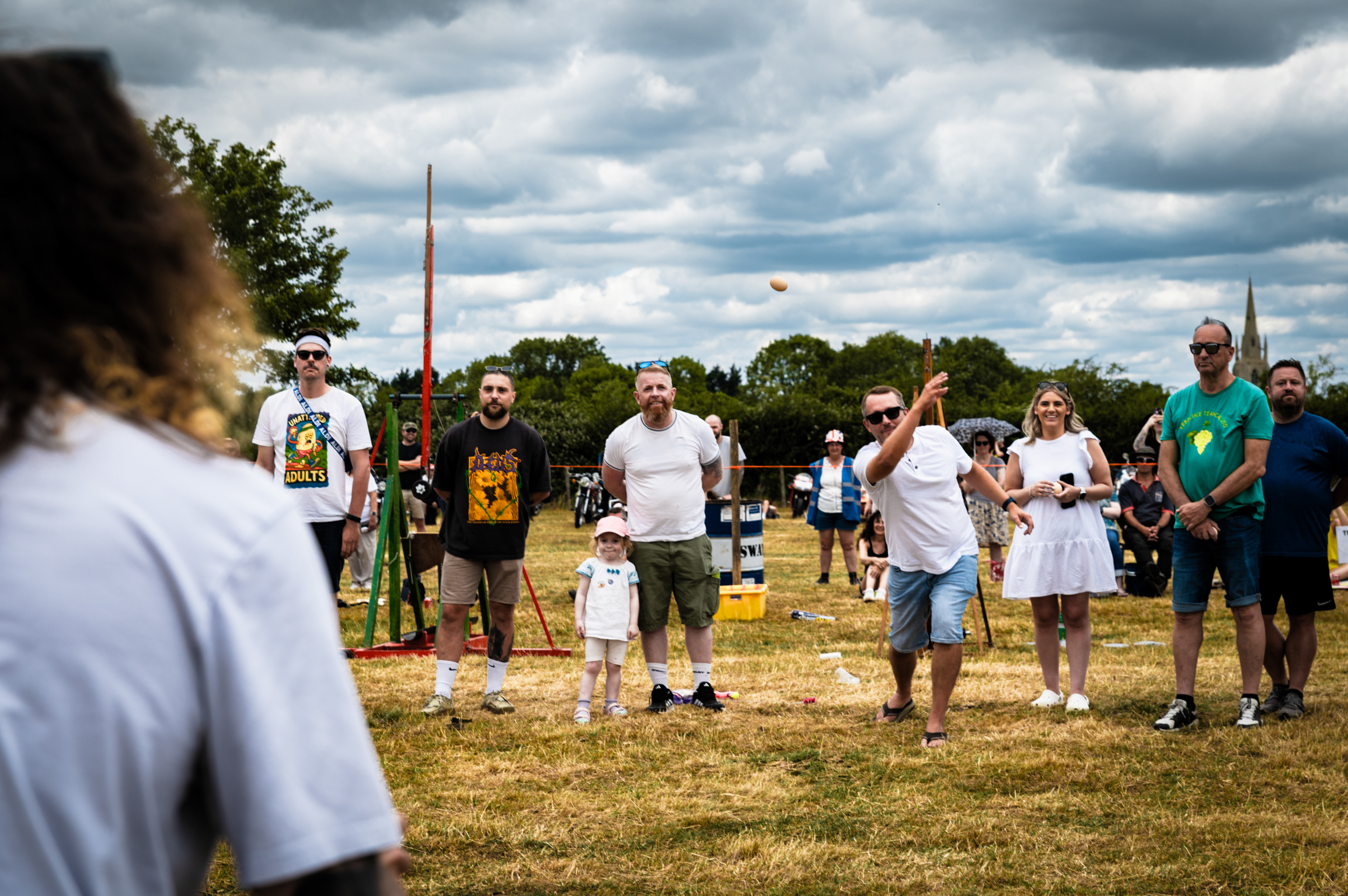 Group of people standing on grass field under cloudy sky, with trees in background and blurred figure in foreground.
