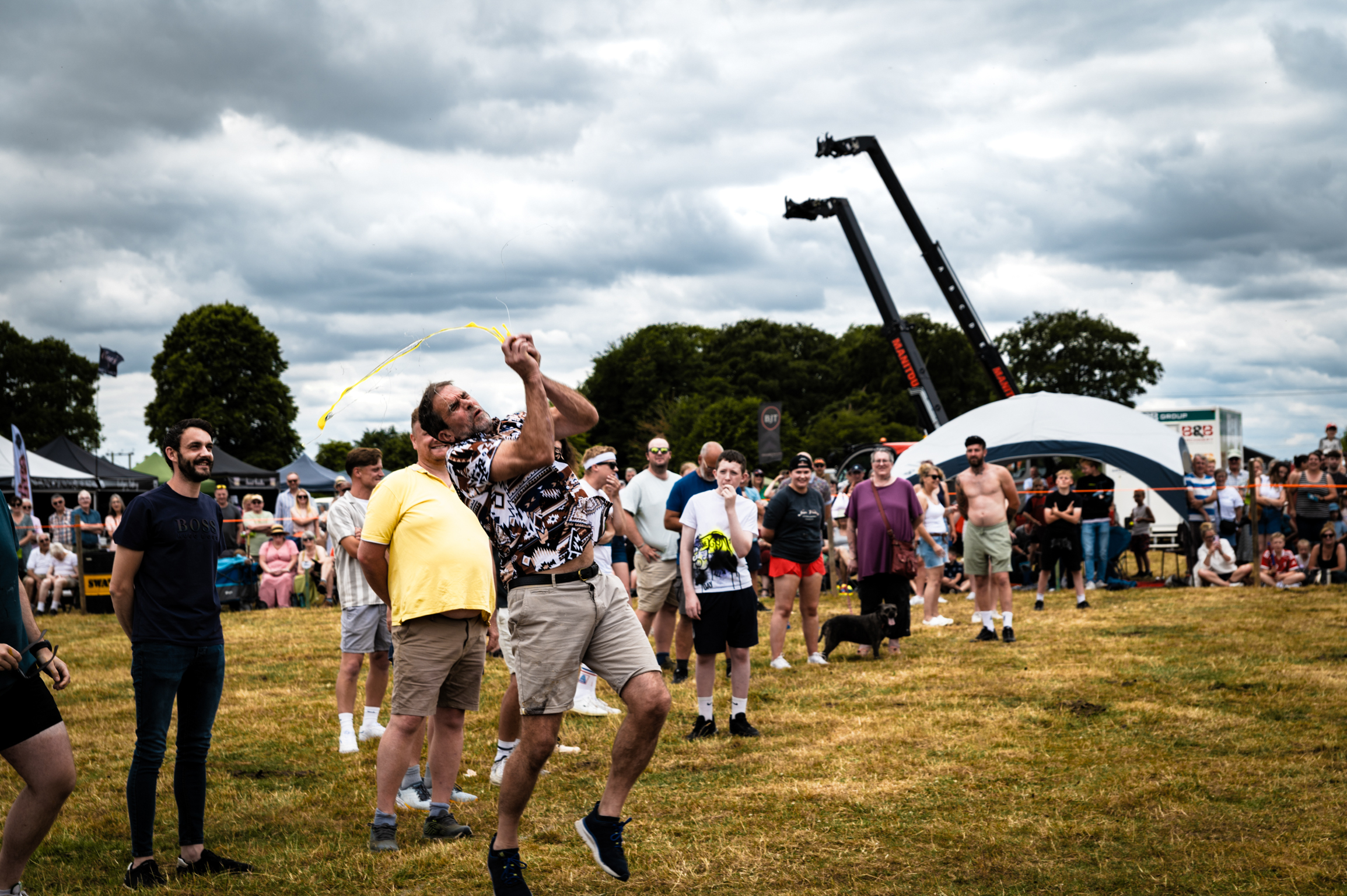 Men competing in hammer throw at outdoor country fair with crowds watching under cloudy sky, green trees in background.